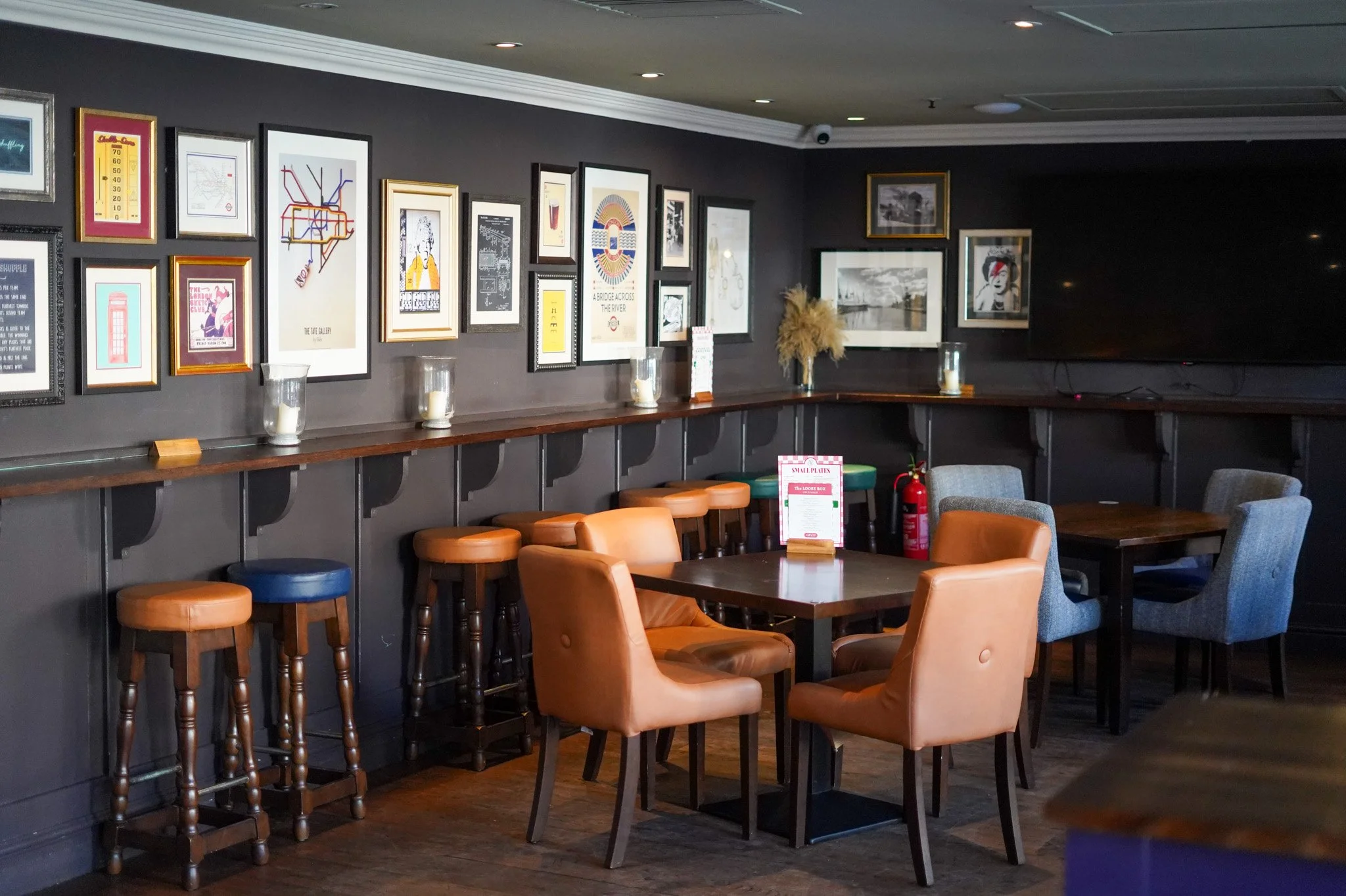 Interior of a cafe or lounge with framed artwork on dark gray walls, wooden tables, and a variety of chairs and stools in tan, blue, and gray colors, with a large flat-screen TV on the wall.