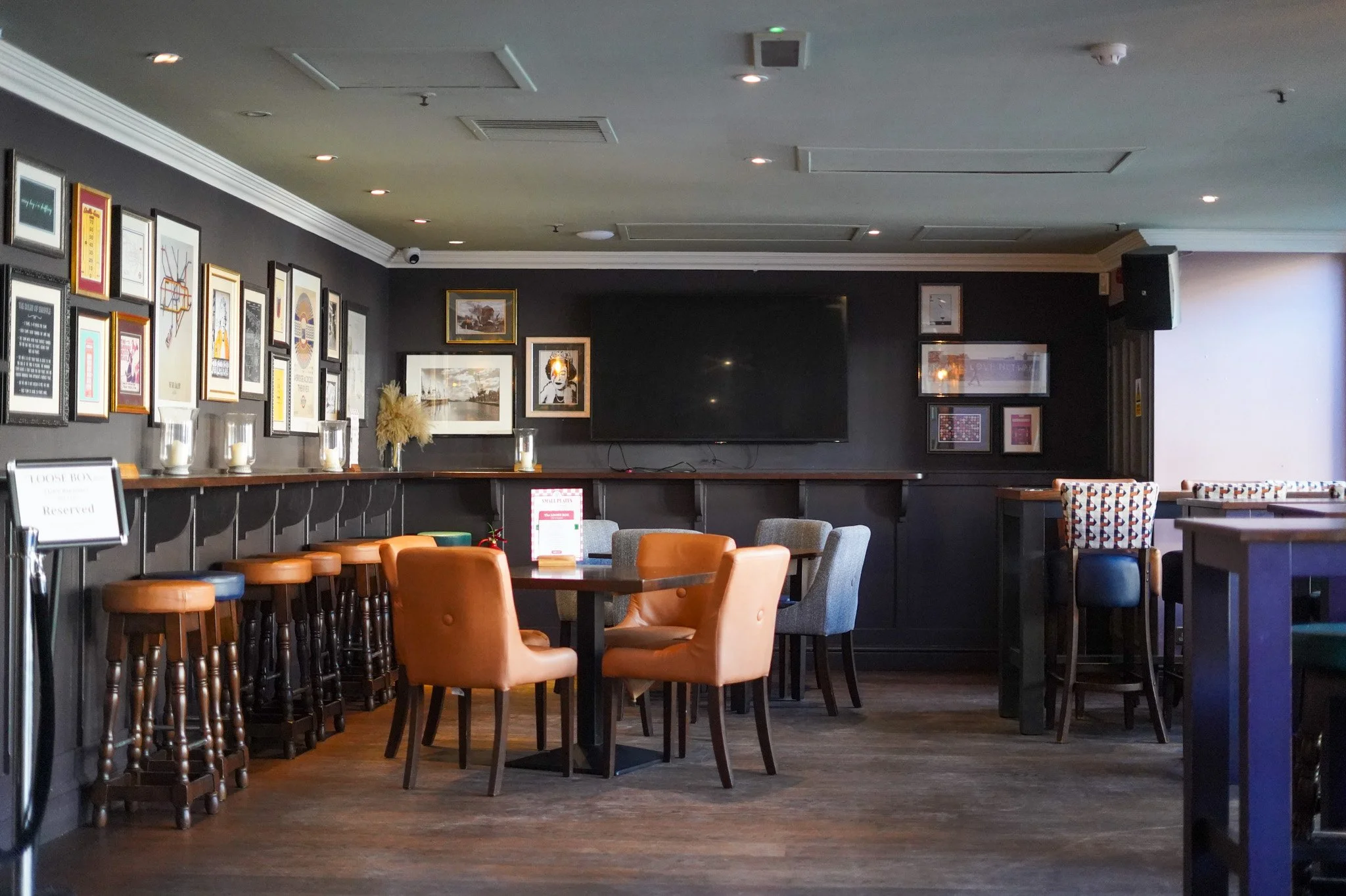 Interior of a cozy bar or restaurant with framed pictures on dark walls, a large TV, tables with chairs in various styles, and a reserved sign on one table.