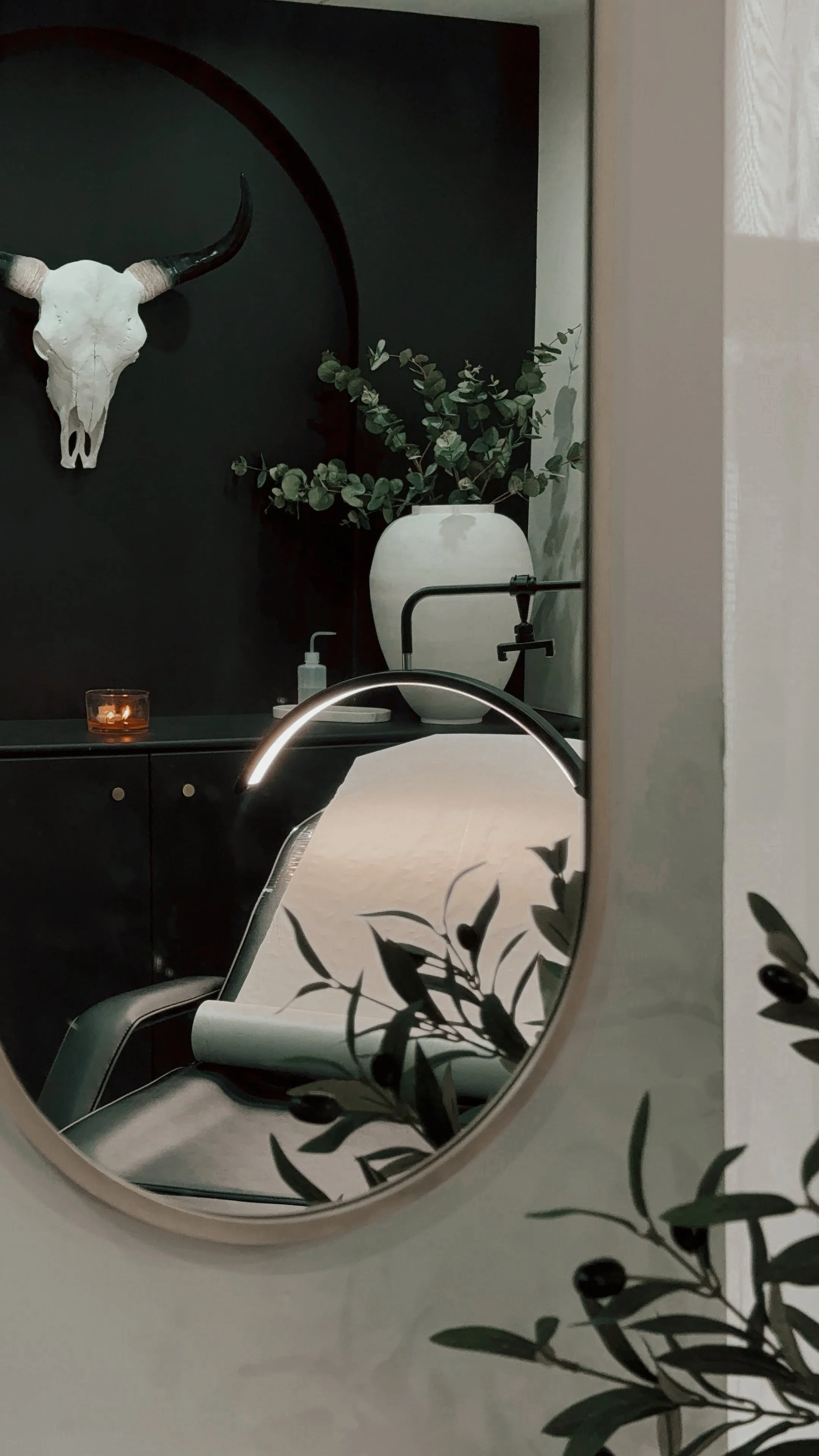 A minimalist room with a decorative bull skull on a black wall, a white vase with greenery, a mirror reflecting a chair, candle, and soap dispenser, and olive branch decor in the foreground.