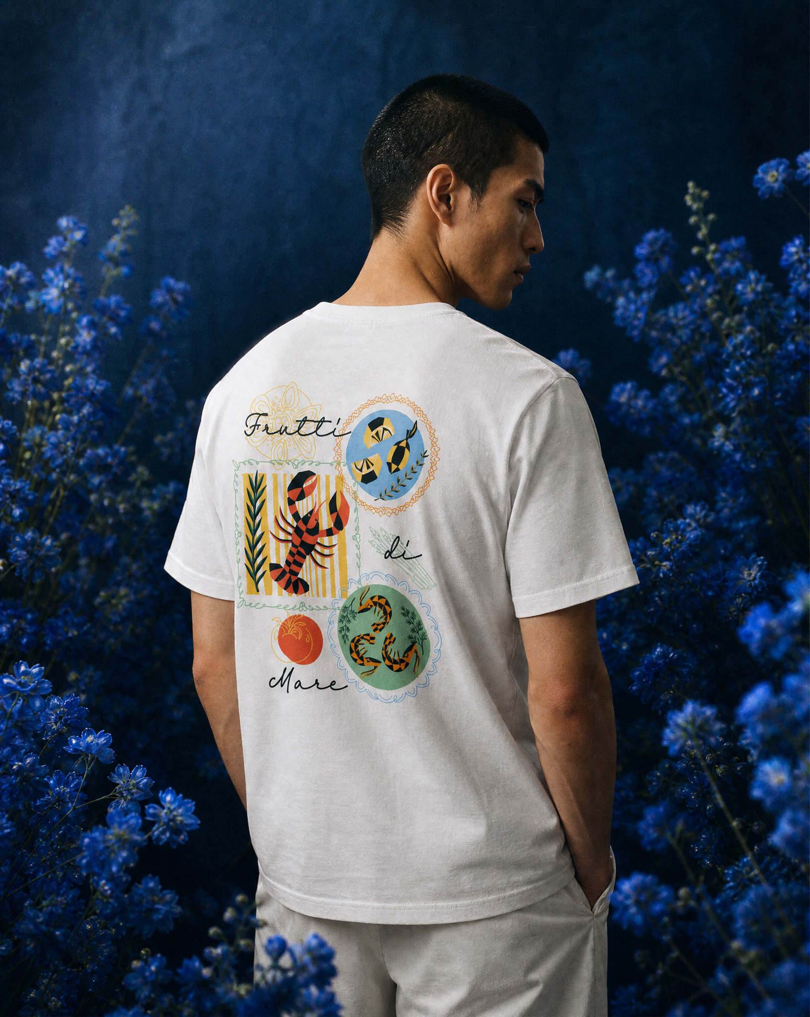 A man with his head turned to the side, wearing a white T-shirt with colorful illustrations and text, standing in front of a dark blue background with blue flowers.