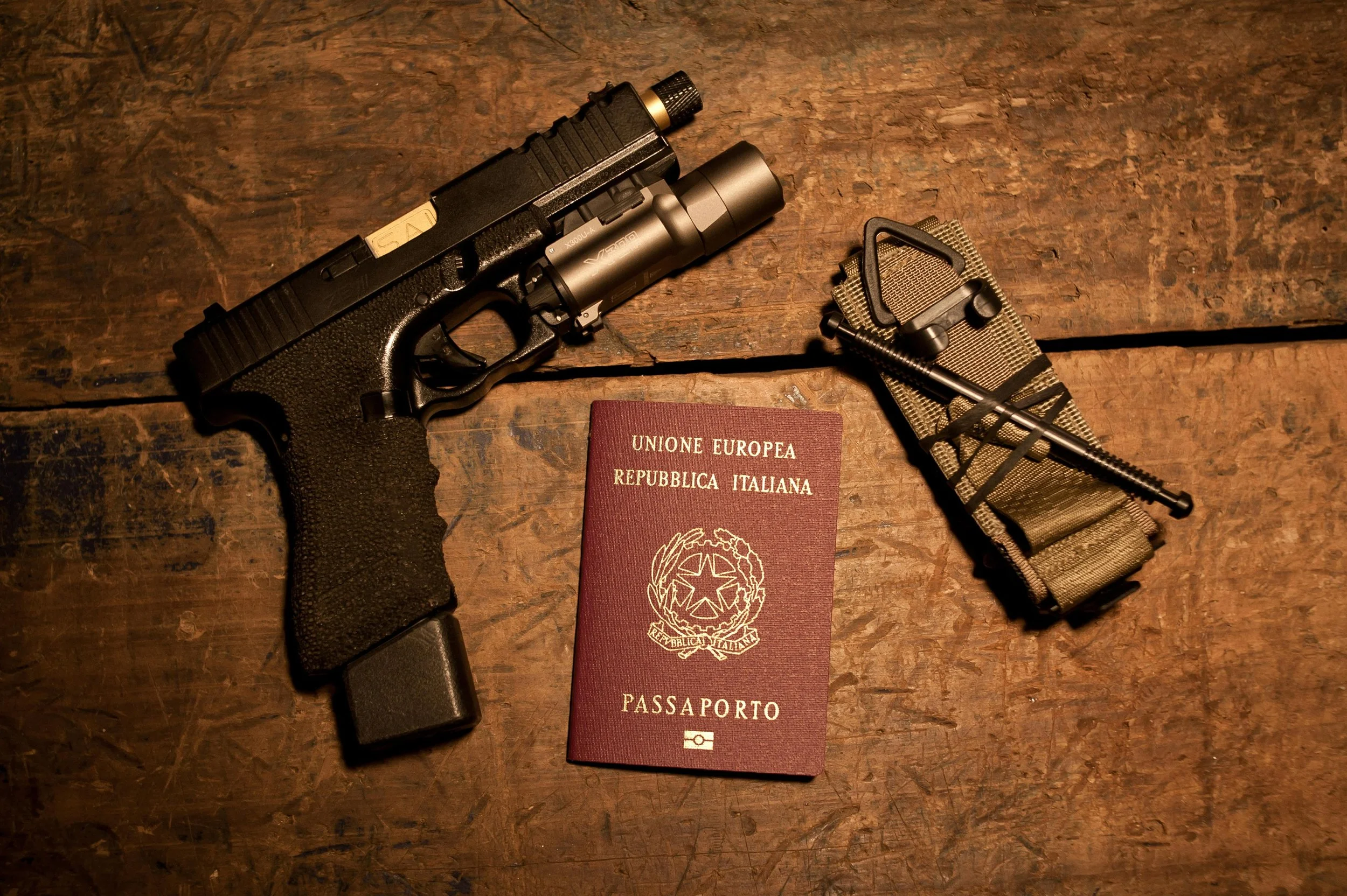 A handgun with a tactical flashlight, a camouflage cover, and a folded European passport on a wooden surface.