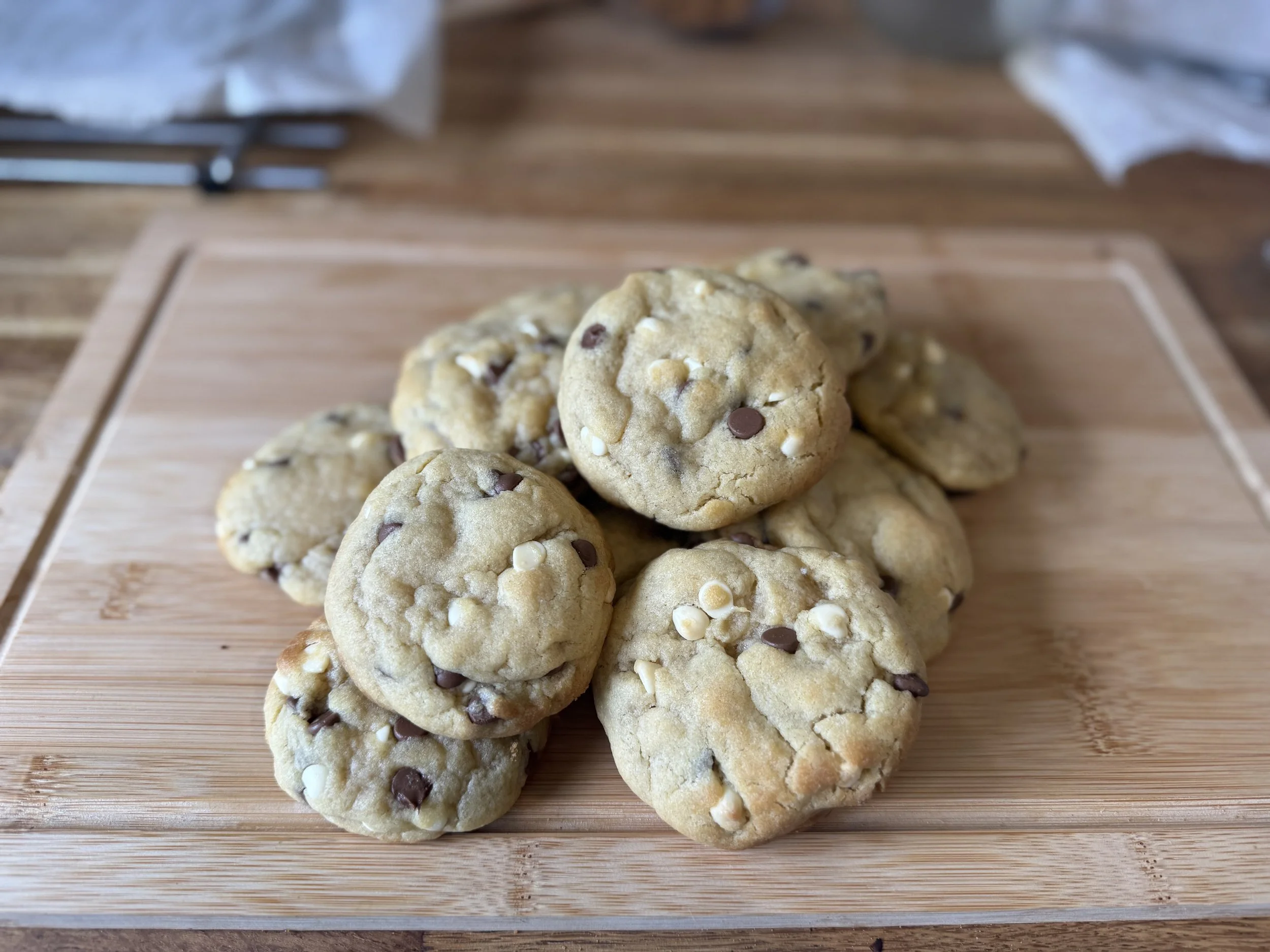 A pile of chocolate chip cookies with white chocolate chips on a wooden cutting board.