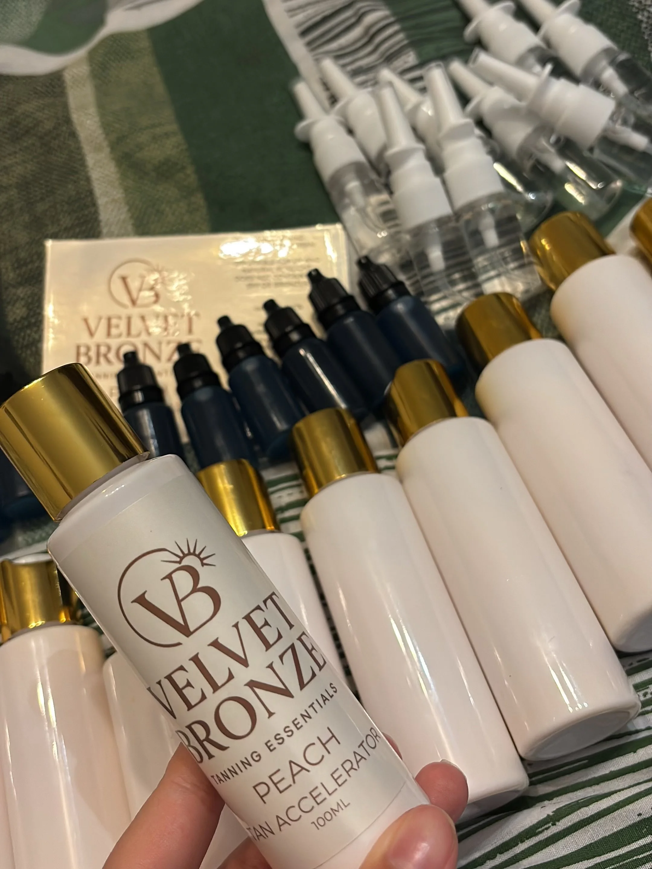 A hand holding a bottle of Velvet Bronze Peach tanning accelerator in front of a display of similar bottles and small spray bottles, all on a green and white striped surface.
