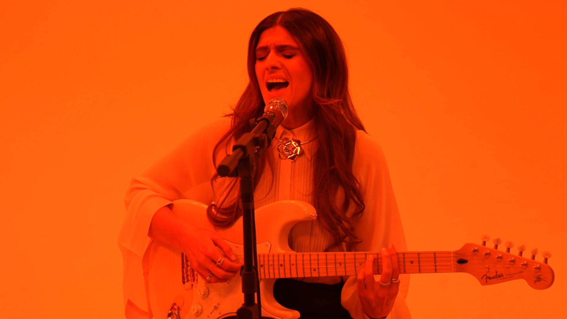 A young woman with long dark hair, wearing a beige blouse, ripped jeans, white sneakers, and socks, is sitting on a pink armchair, talking on a vintage pink telephone, smiling, with pink curtains in the background.