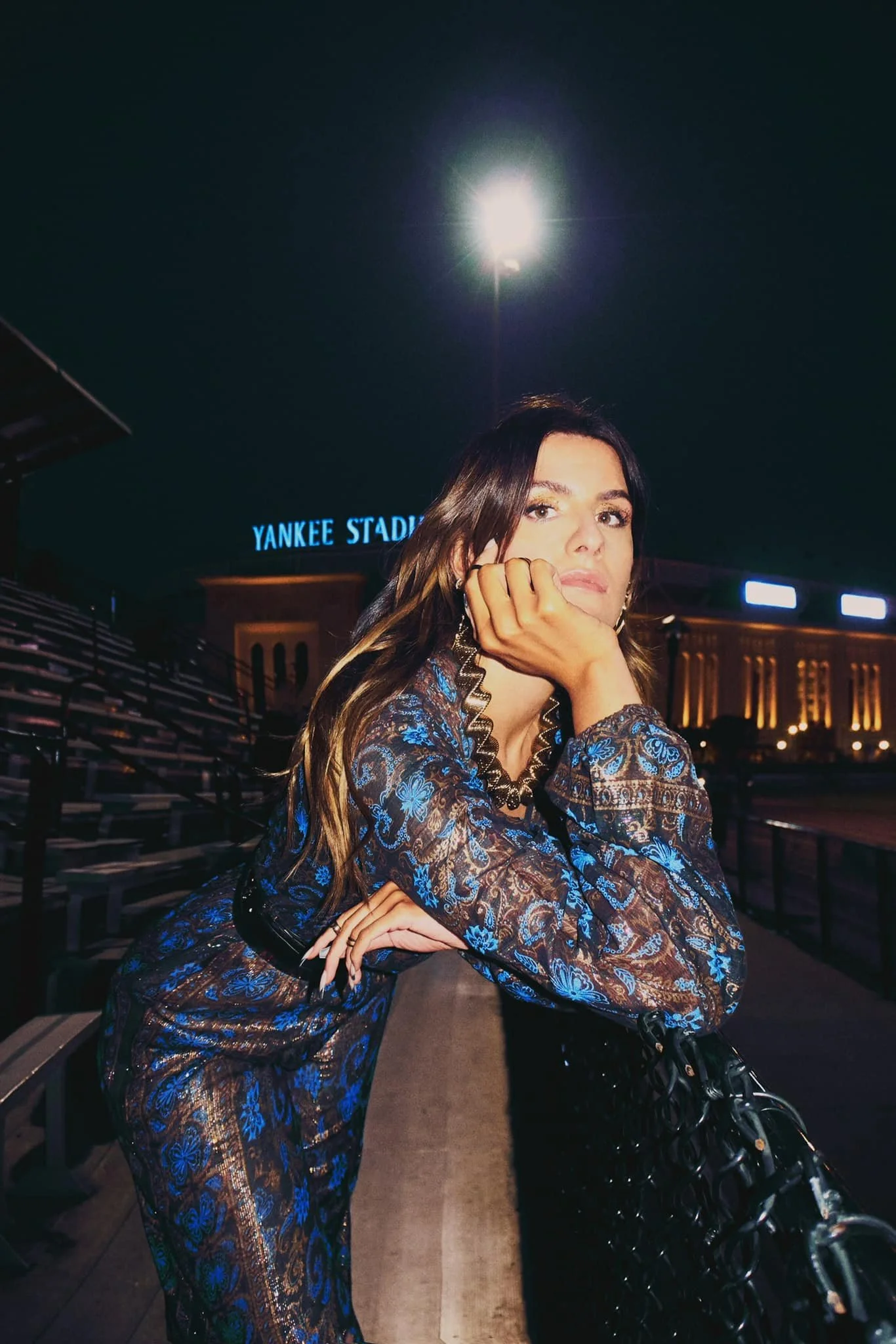 A woman with long dark hair is sitting on a bench at night outside Yankee Stadium, resting her head on her hand and looking at the camera. She is wearing a patterned blue and brown outfit and has a black purse on her lap. The stadium's illuminated sign and seating area are visible in the background.