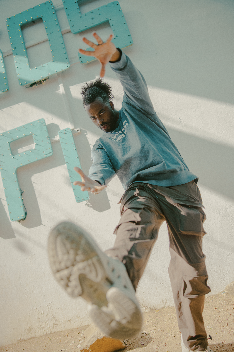 A man with braided hair wearing a black hoodie and black athletic pants, standing in a dimly lit urban parking garage, adjusting the hood on his head.