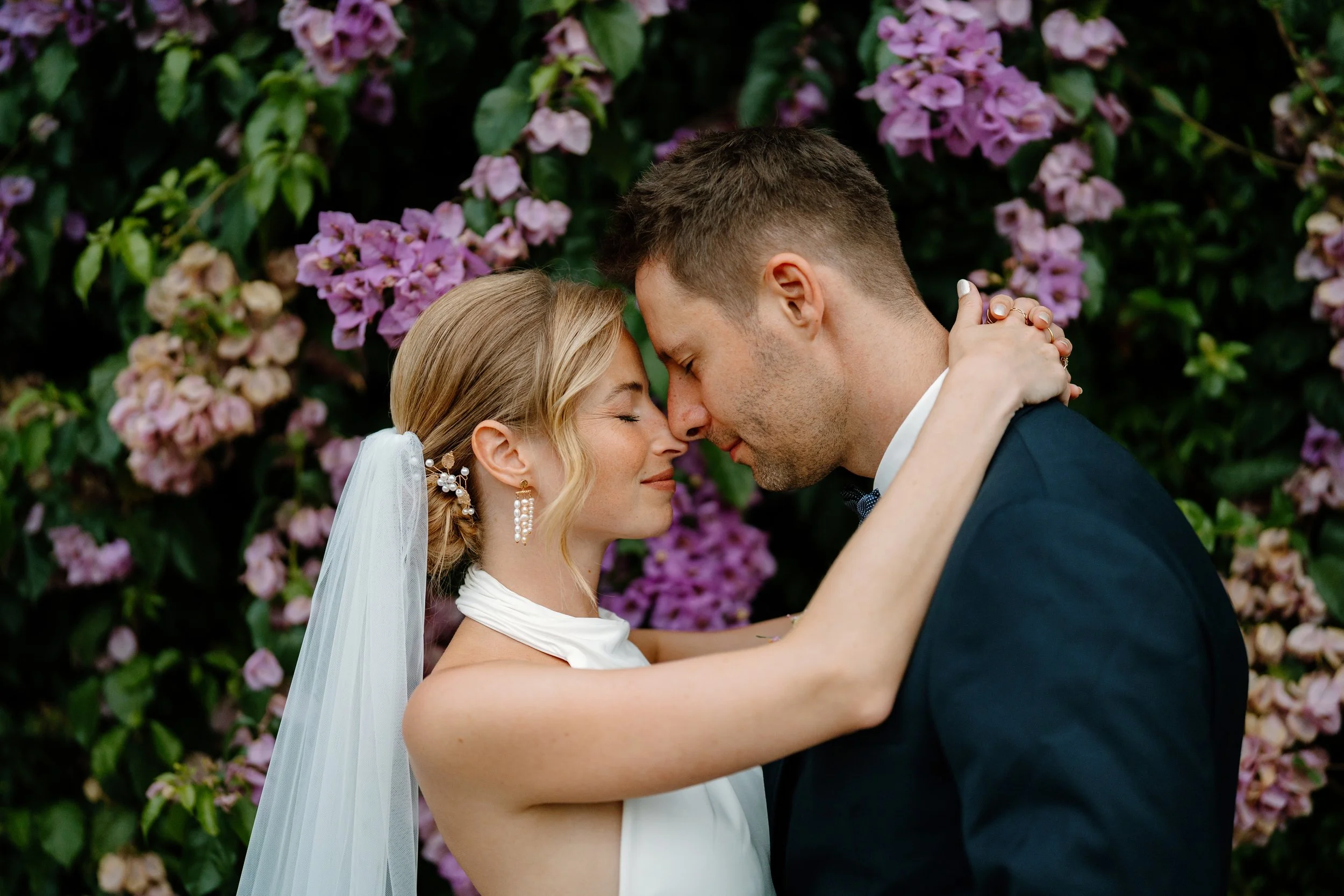 A bride and groom embrace with foreheads touching, eyes closed, standing in front of blooming purple flowers.