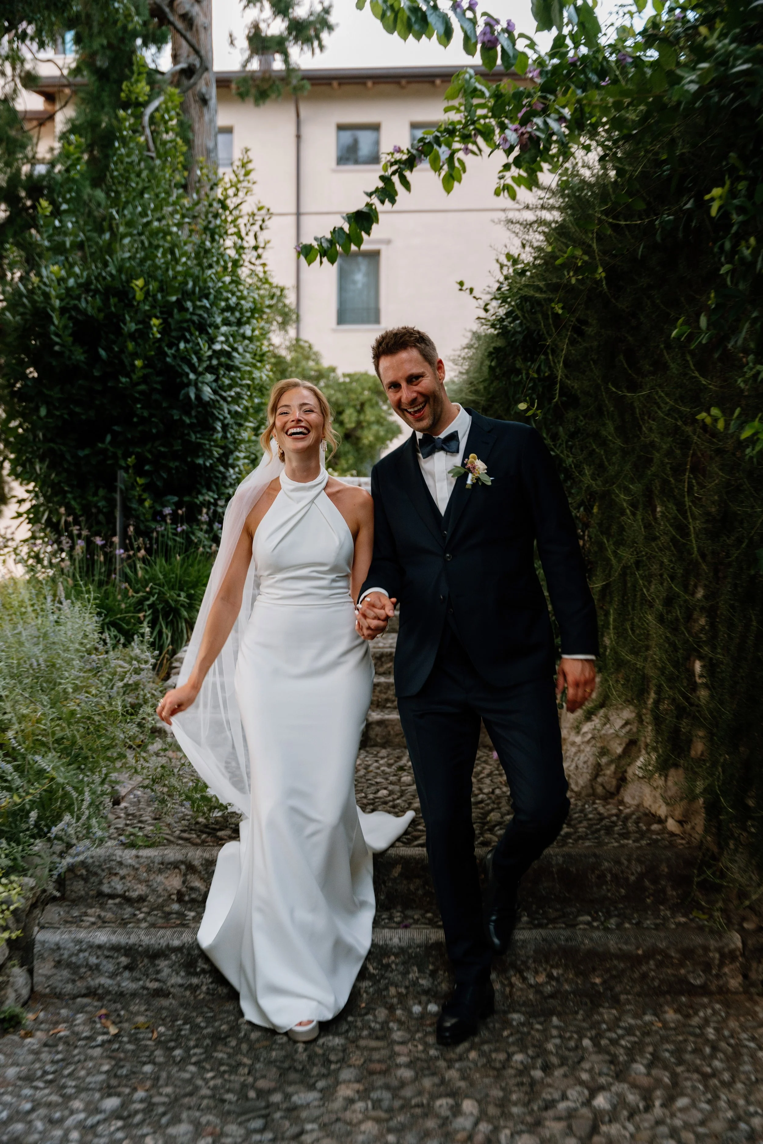 A bride and groom walking together, smiling and holding hands, outdoors on stone steps surrounded by greenery, during their wedding day.