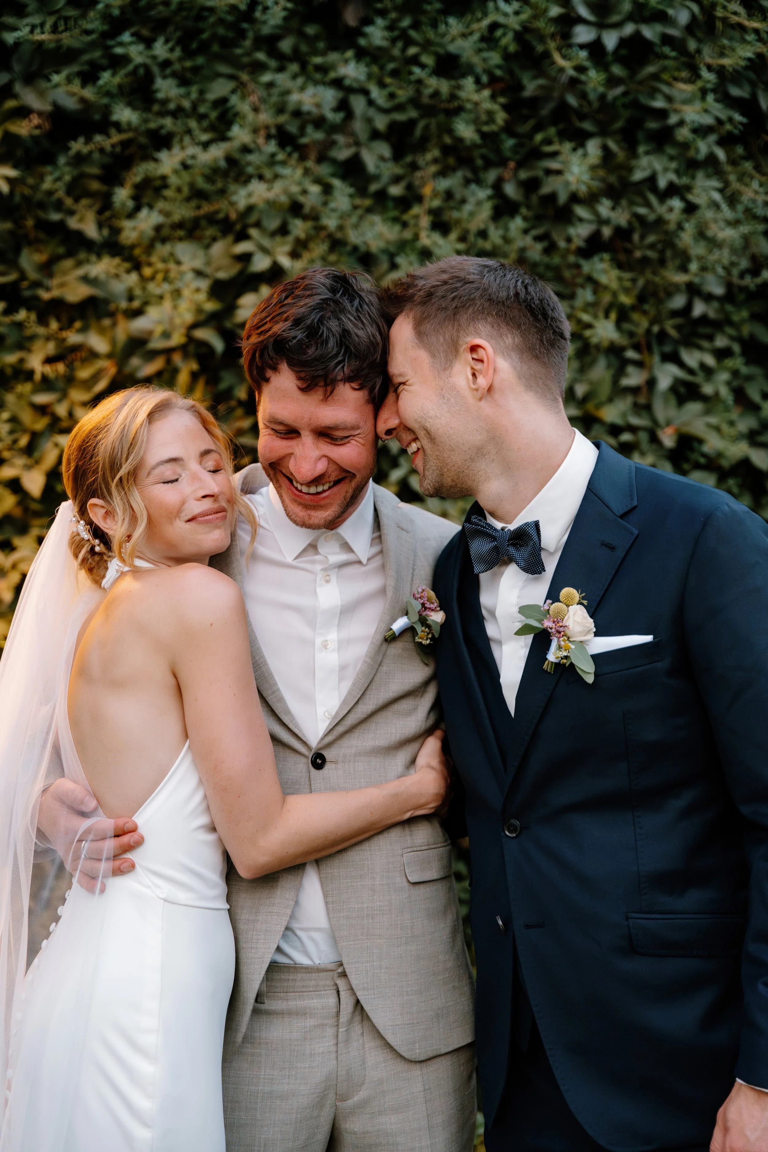 A bride and two grooms, all smiling and sharing a joyful moment at their wedding, standing close together in front of a leafy background.