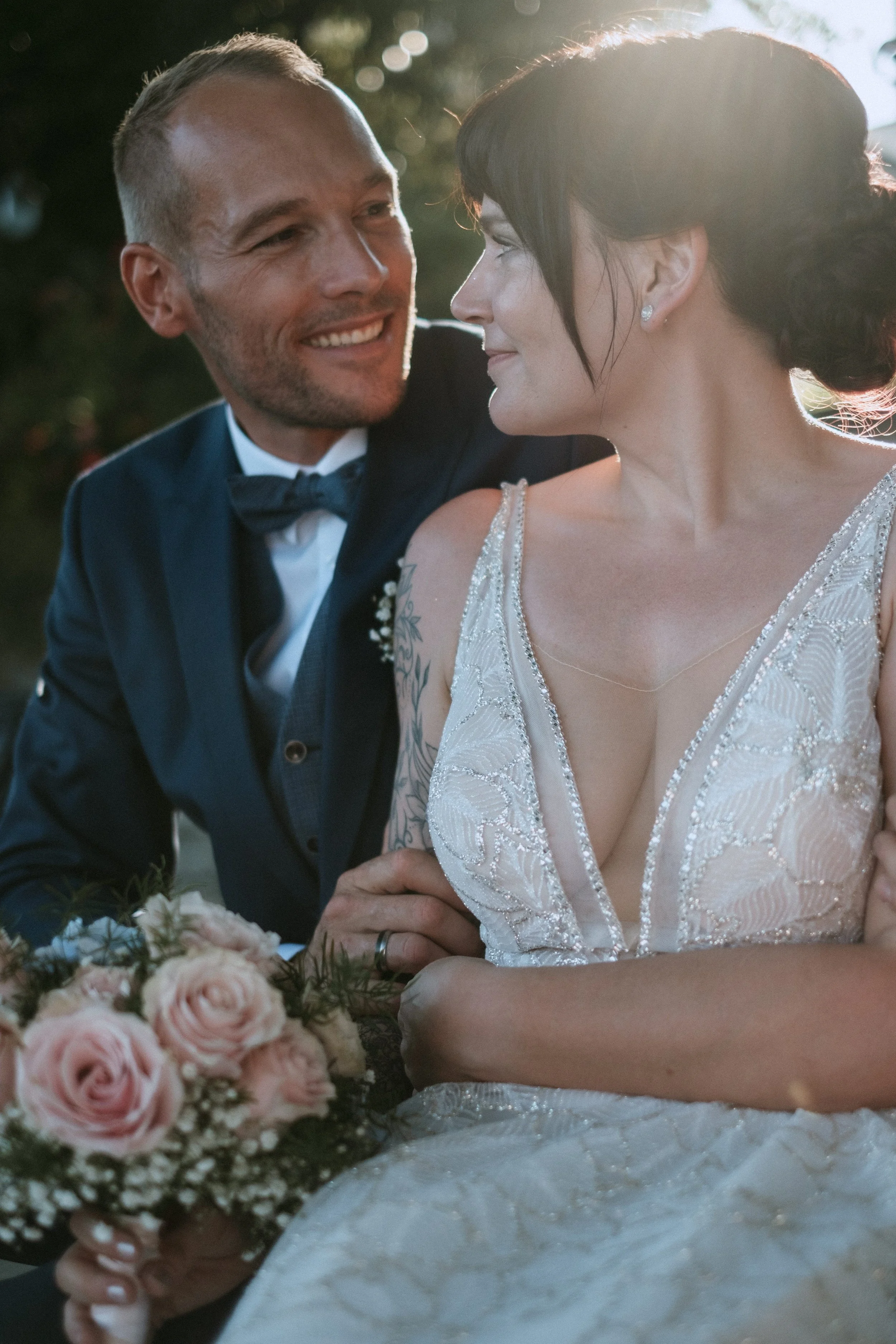 A couple dressed in wedding attire, the man in a dark suit and bow tie, and the woman in a white lace wedding gown with a deep neckline, sharing an intimate moment outdoors with sunlight behind them, holding a bouquet of pink and white roses.