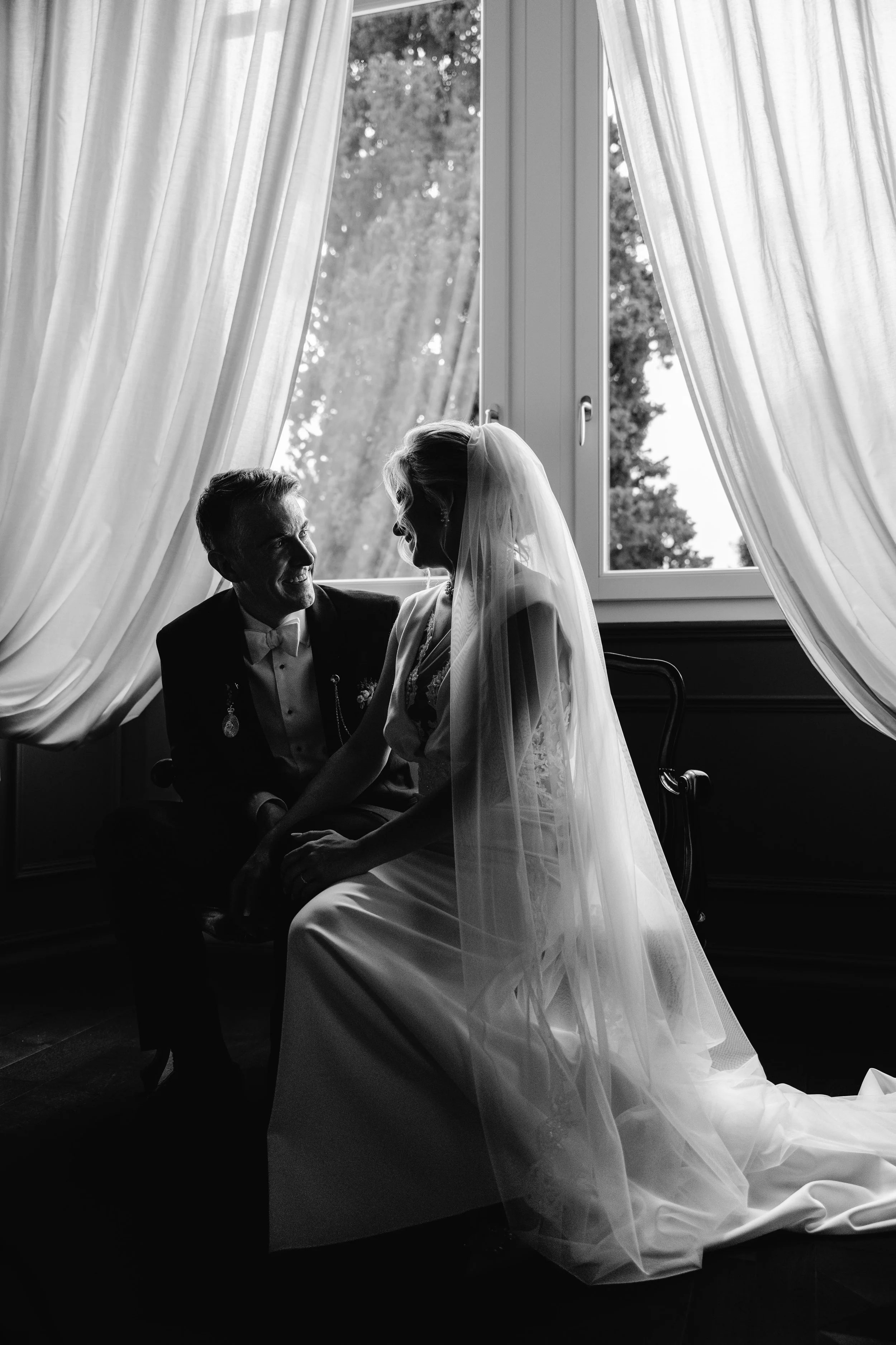 Black and white photo of a bride and groom sitting near a window with curtains, facing each other and smiling.