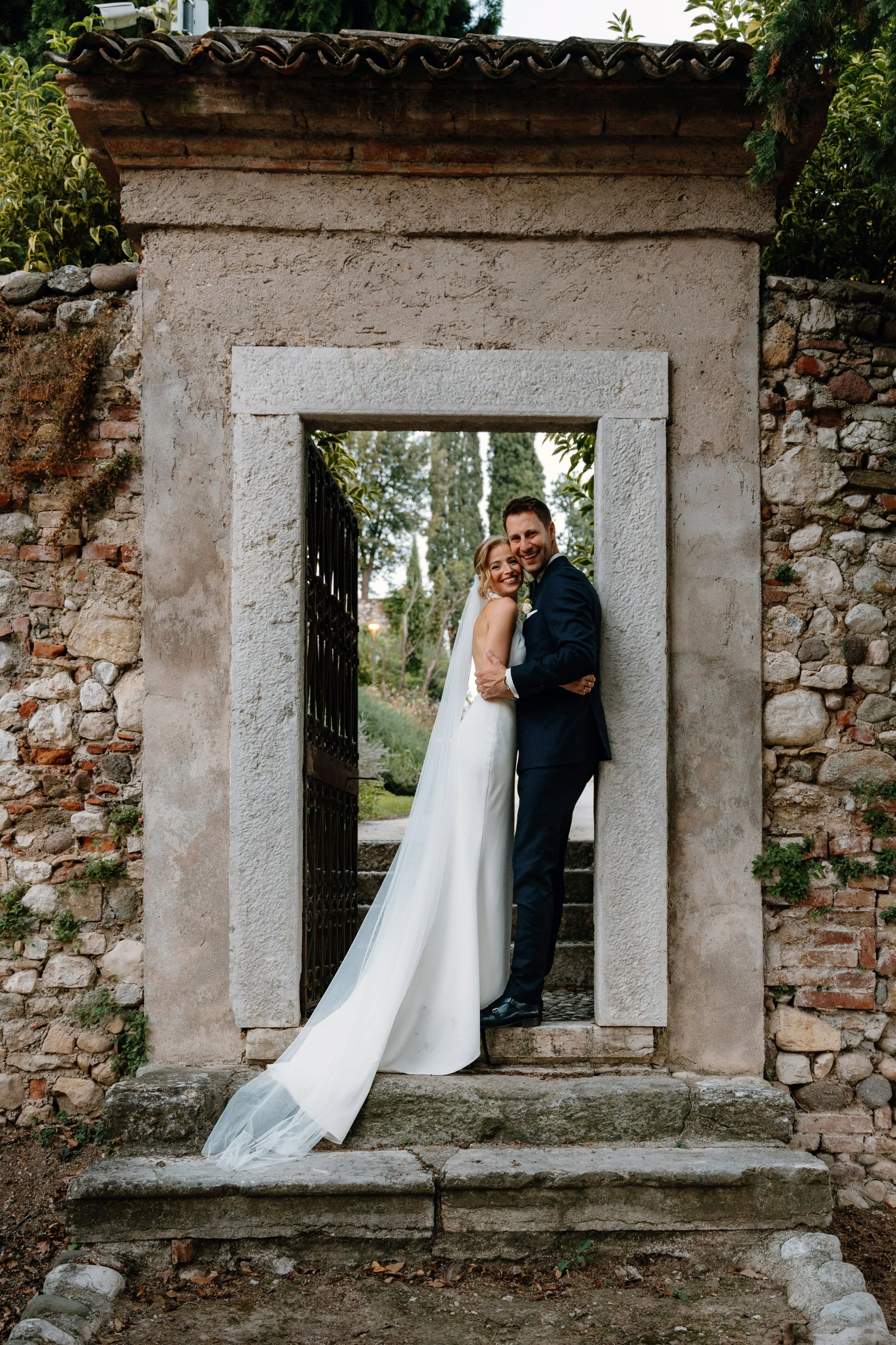 A bride and groom smiling and hugging in a wedding photo, standing in a stone and brick doorway outdoor setting.