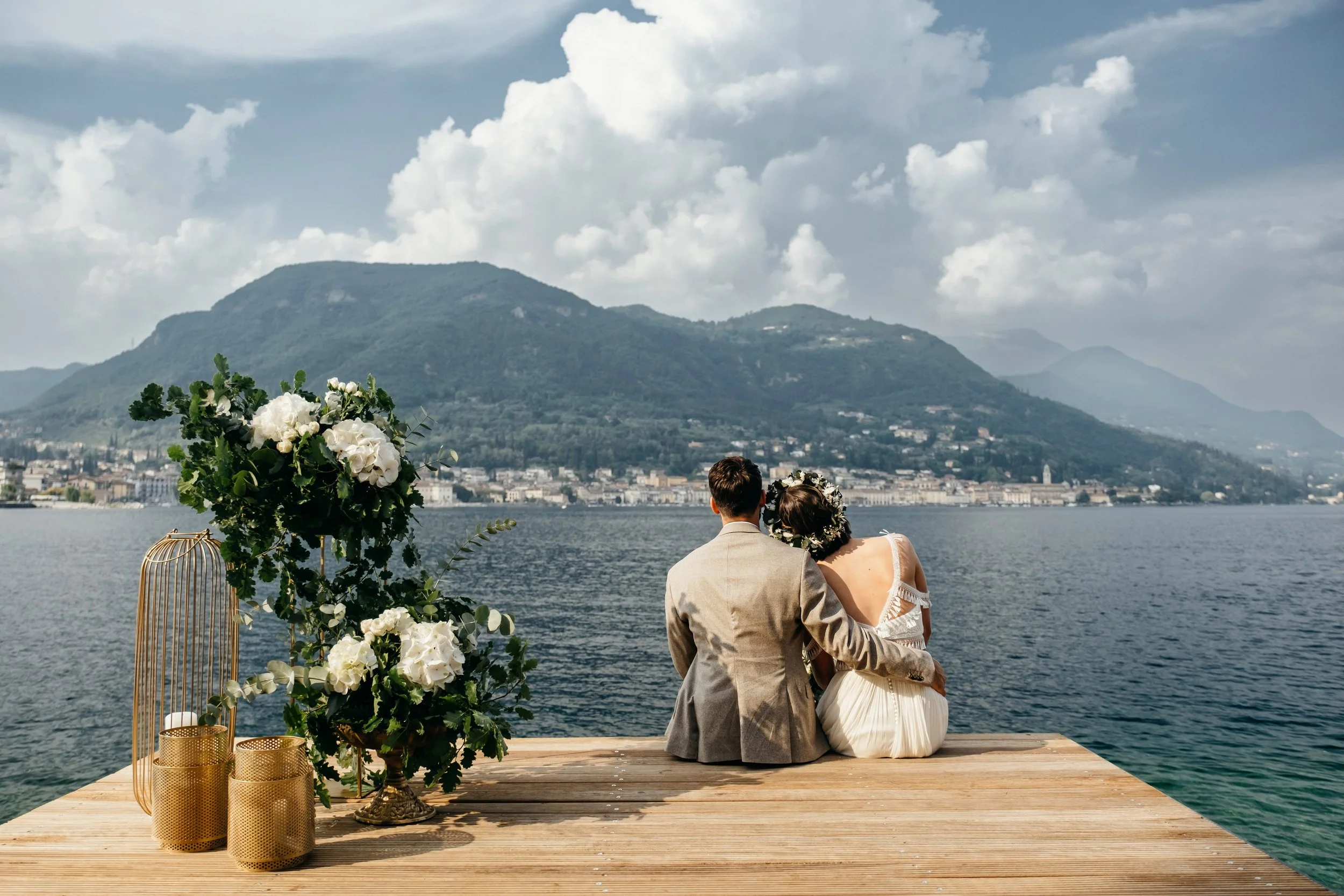 A couple sitting on a wooden dock by a lake, with mountains and a cloudy sky in the background; the woman is wearing a wedding dress with off-the-shoulder sleeves and a floral crown, and the man is in a beige suit. They are facing away from the camera, holding each other, with floral arrangements and candle holders nearby.