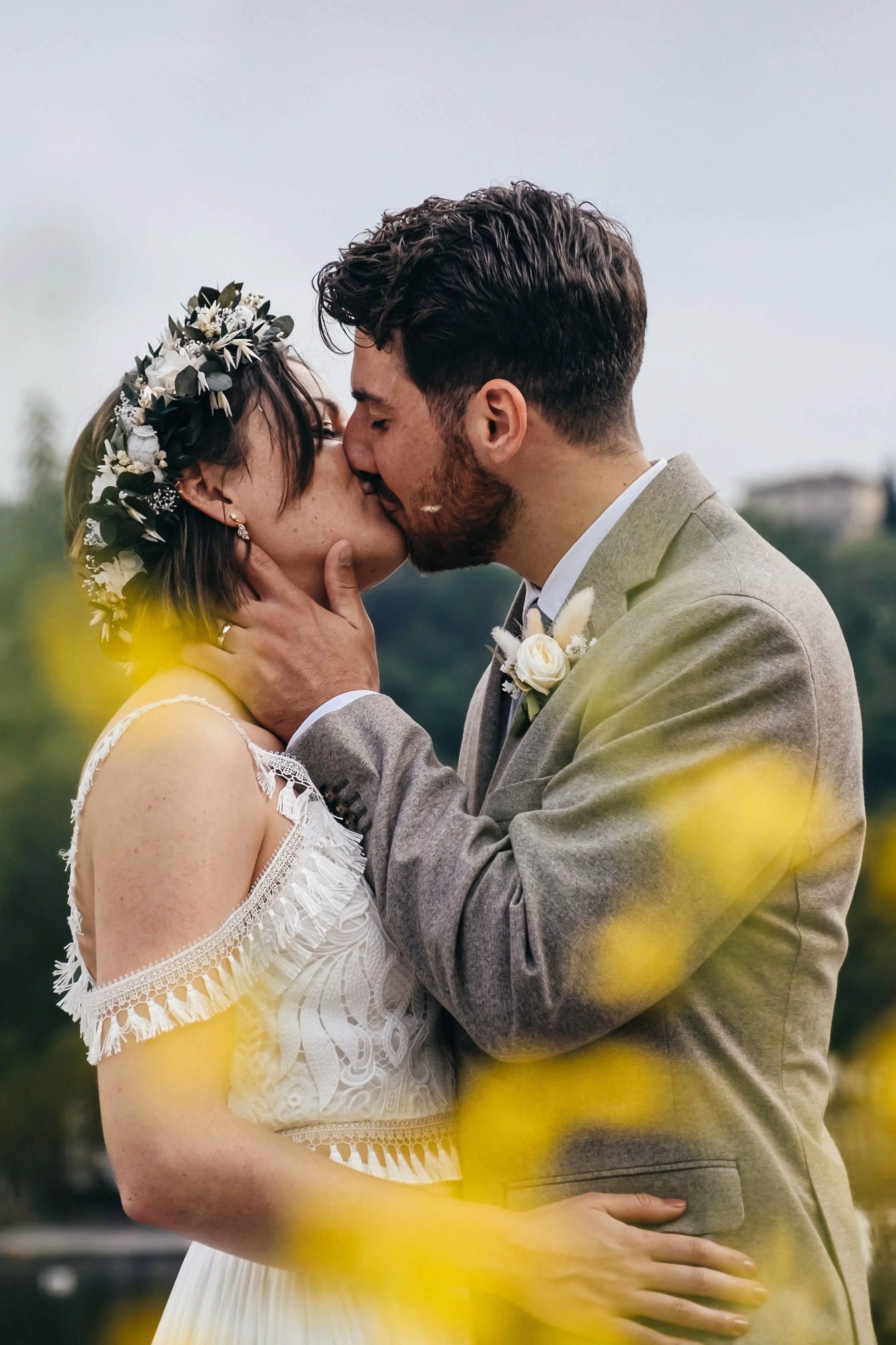 A bride and groom kissing outdoors, surrounded by yellow flowers, with greenery and distant buildings in the background.