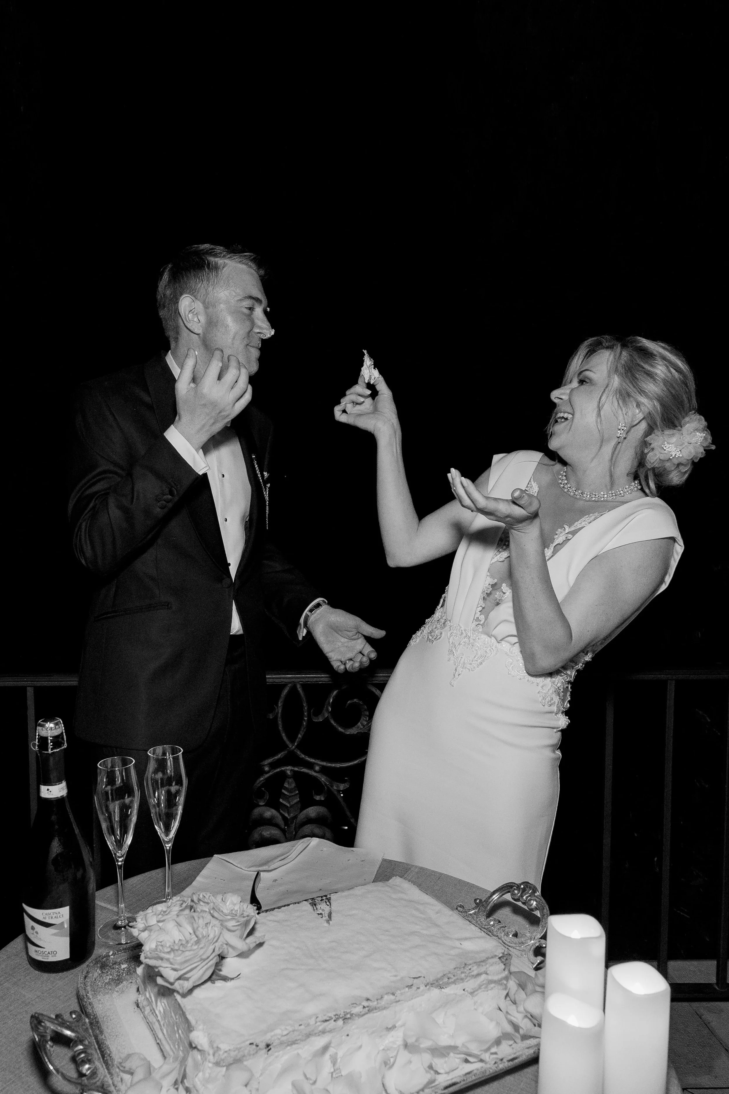 A bride and groom celebrating at their wedding reception, standing beside a cake with candles and flowers, in a black and white photograph.
