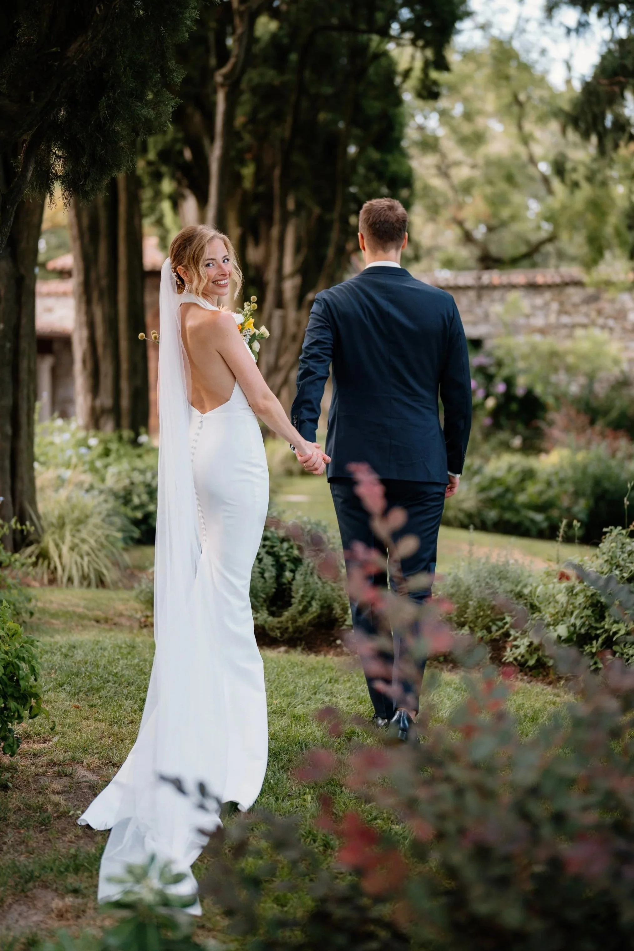 A bride and groom holding hands and walking through a garden, with the bride smiling at the camera.