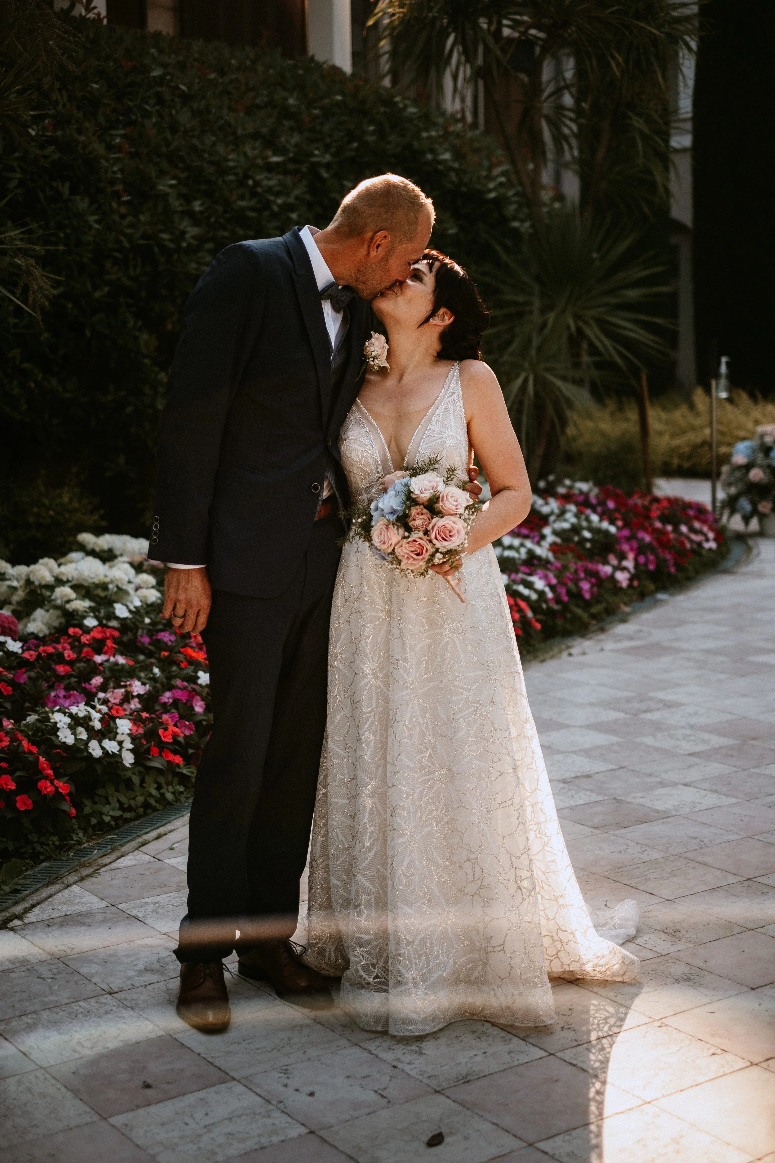 A bride and groom sharing a kiss on their wedding day outdoors, with the bride holding a bouquet of pink roses and blue flowers, and the groom dressed in a black tuxedo, standing on a stone-paved pathway lined with colorful flowers and greenery.
