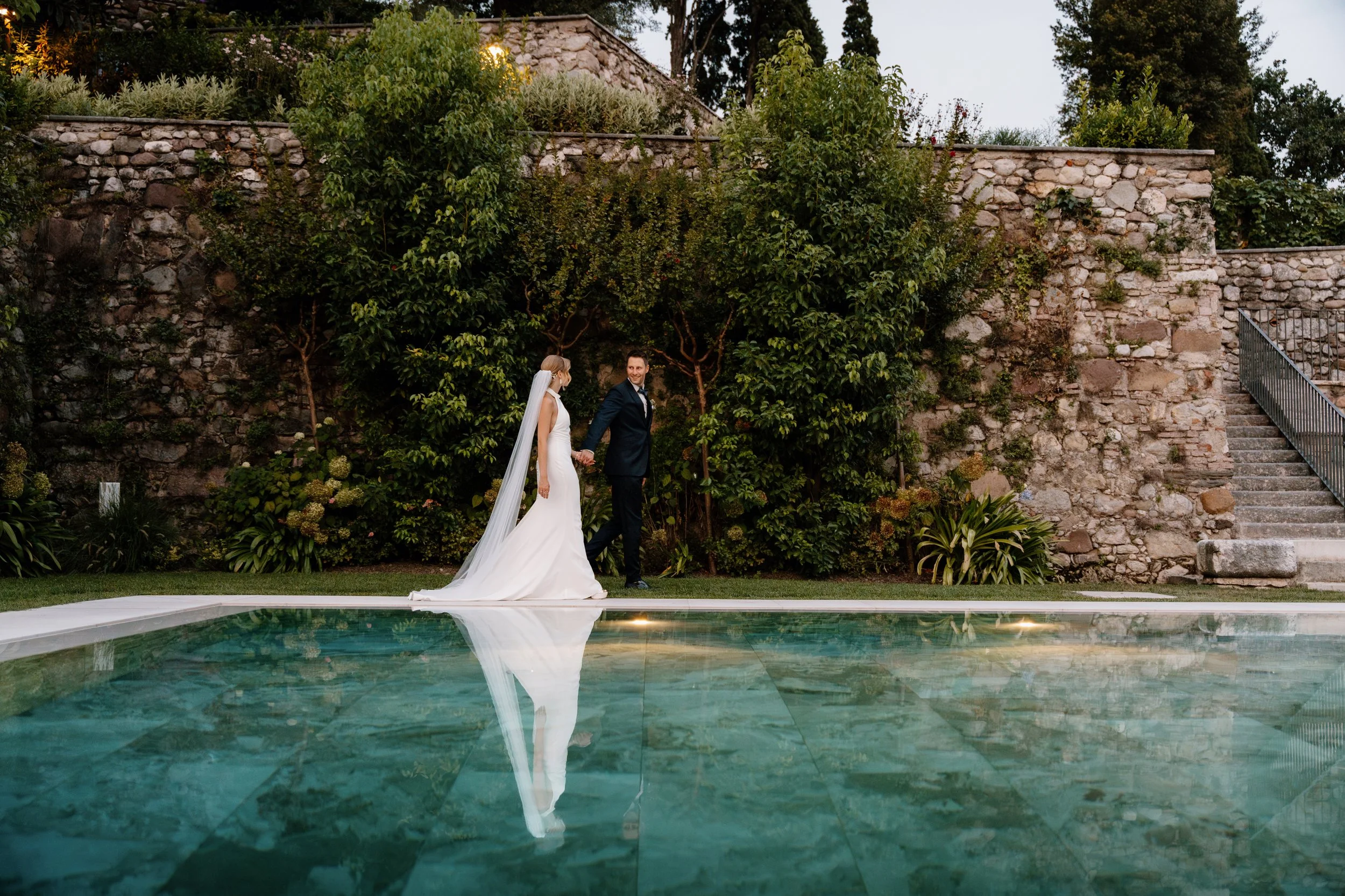 A bride and groom holding hands next to a swimming pool, with lush greenery and a stone wall in the background.