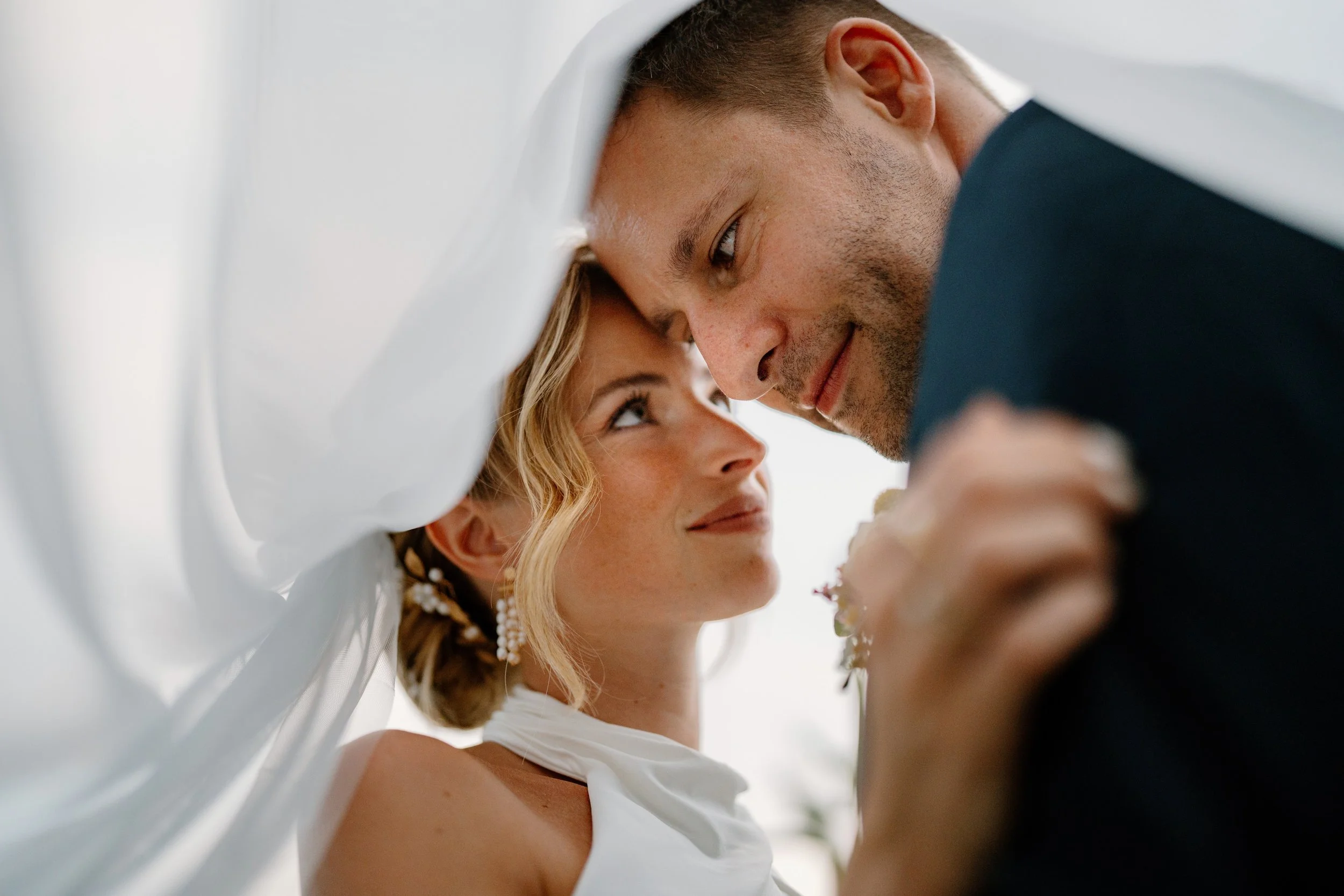 Close-up of a bride and groom under a flowing white veil on their wedding day, looking into each other's eyes with fond expressions.