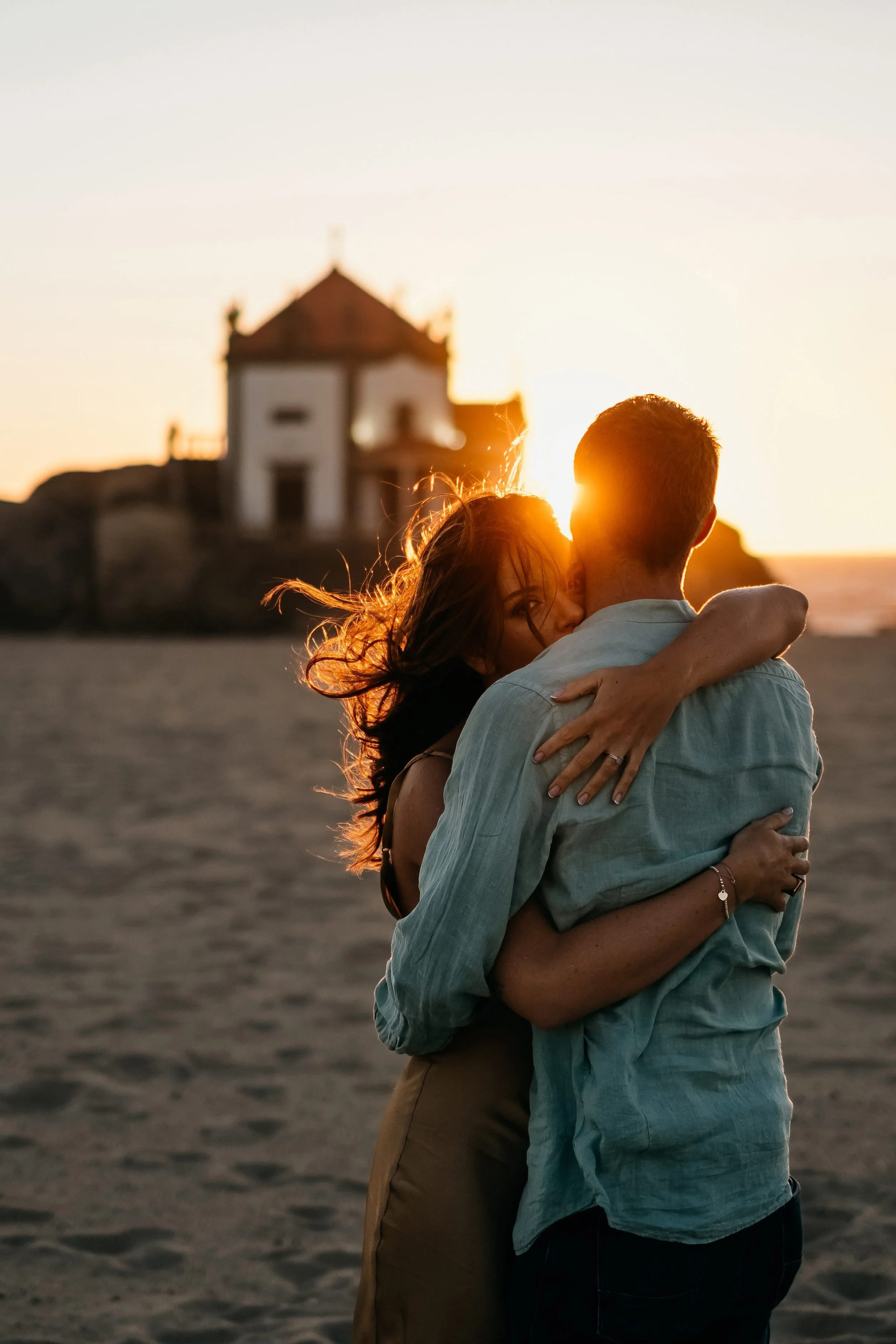 A couple embracing on the beach during sunset with a small house on a hill in the background.