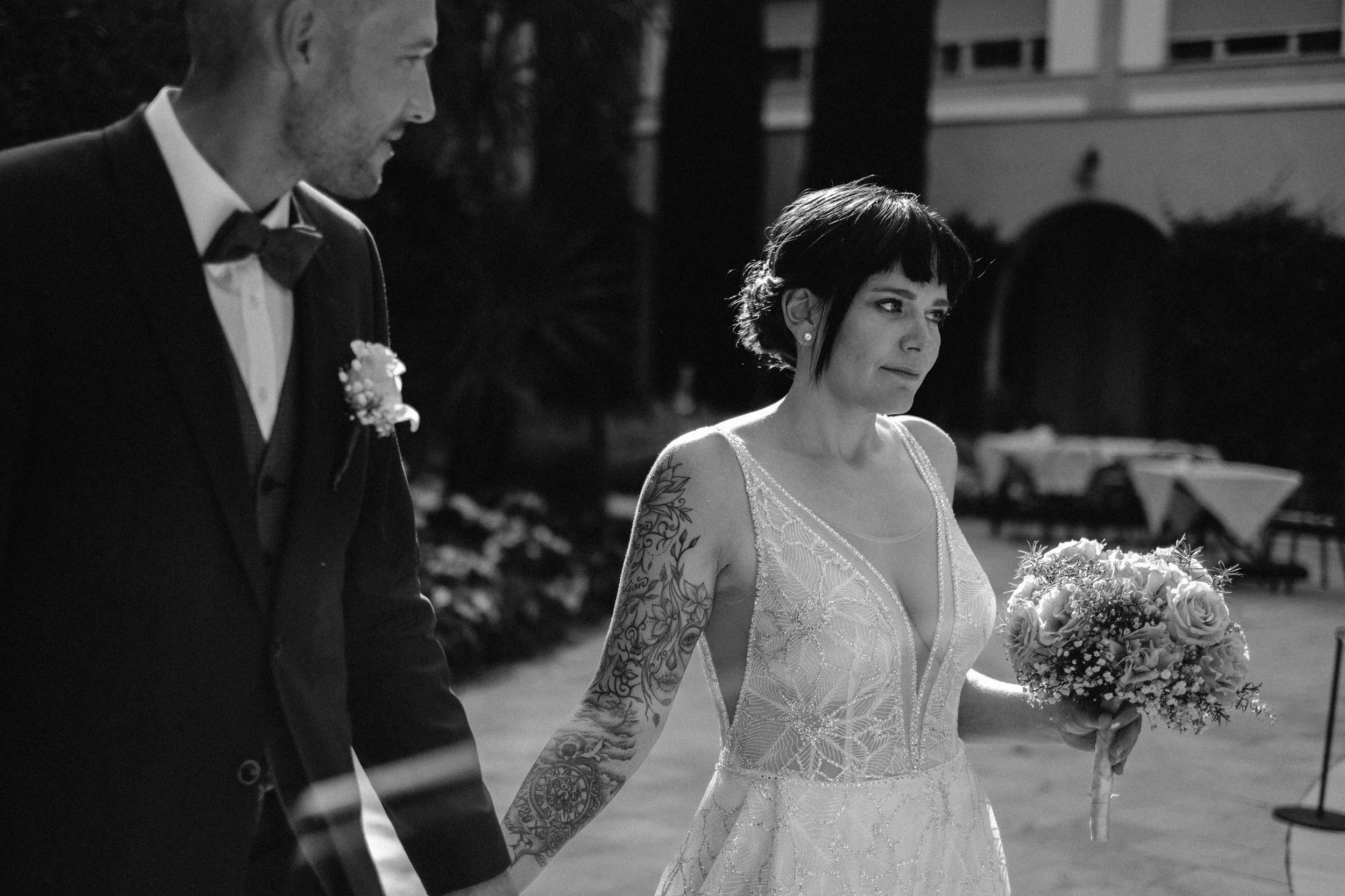 Black and white photo of a bride and groom holding hands outdoors in a wedding setting, with bride holding a bouquet of flowers.