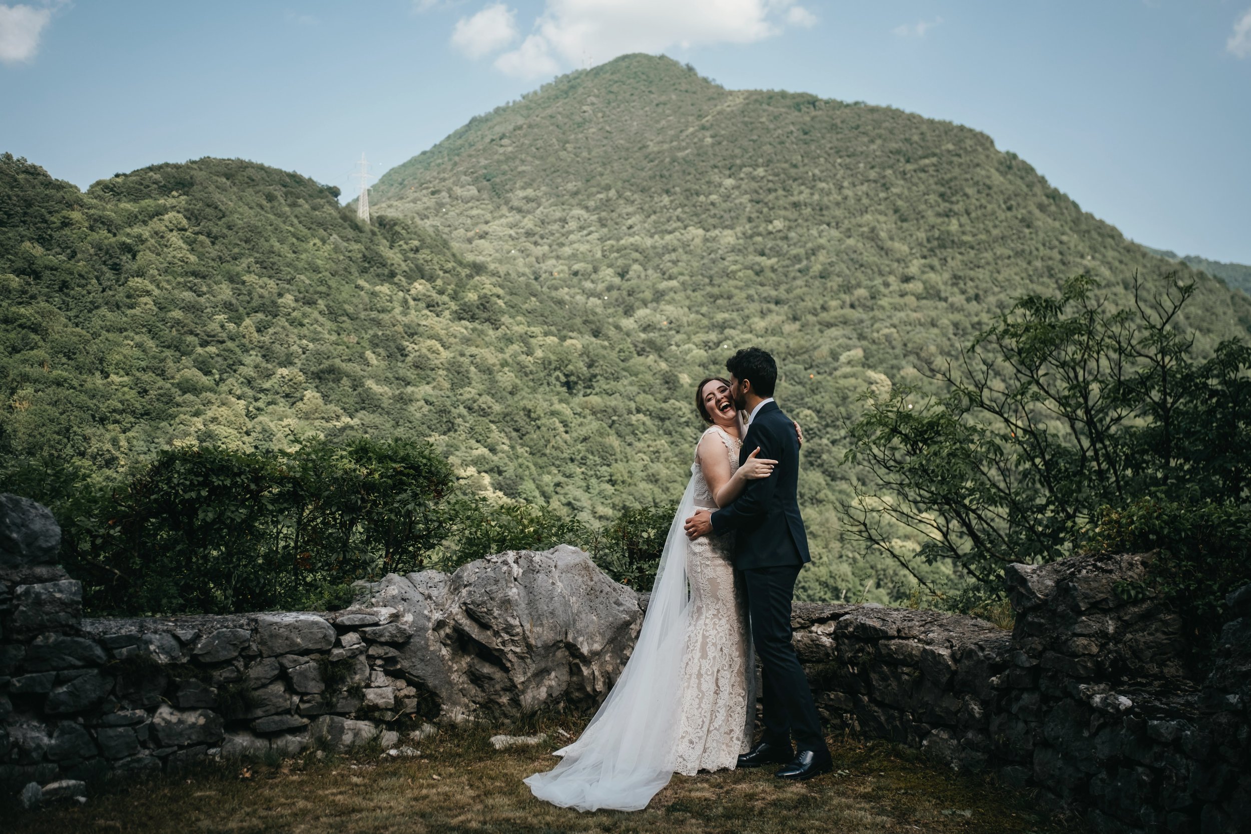 A bride and groom hugging and laughing in front of a lush green mountain landscape during a wedding photoshoot.