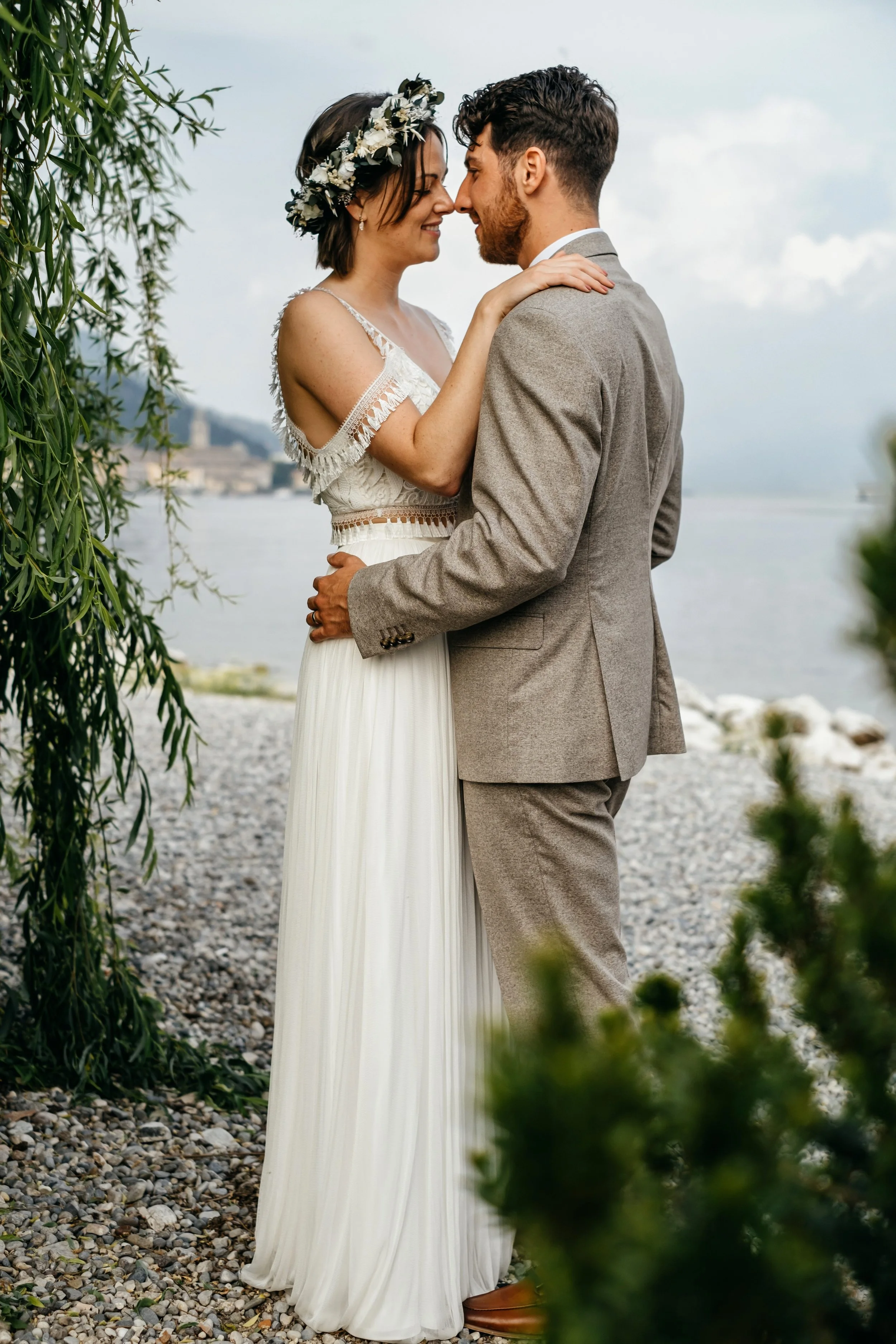 A bride and groom sharing a tender moment outdoors near a body of water. The bride is wearing a white dress with lace details and a floral headband, and the groom is in a gray suit. They are holding each other close, smiling softly as they gaze into each other's eyes.