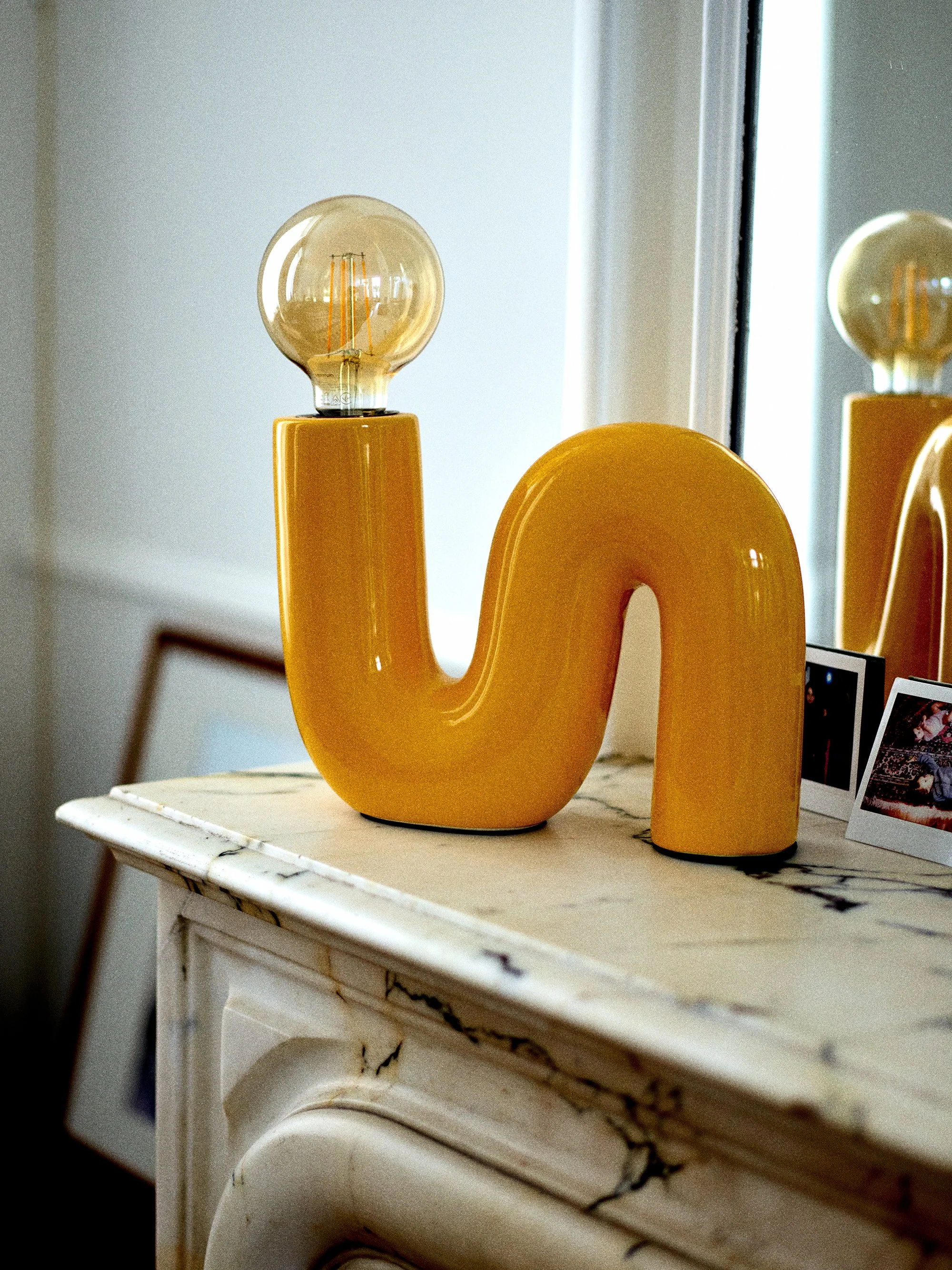 Une lampe décorative en forme de la lettre 'J' de couleur jaune, avec une ampoule à filament en haut, posée sur une table en marbre blanc, près d'un miroir. photographié par un photographe spécialisé en architecture d'intérieur à Lille.