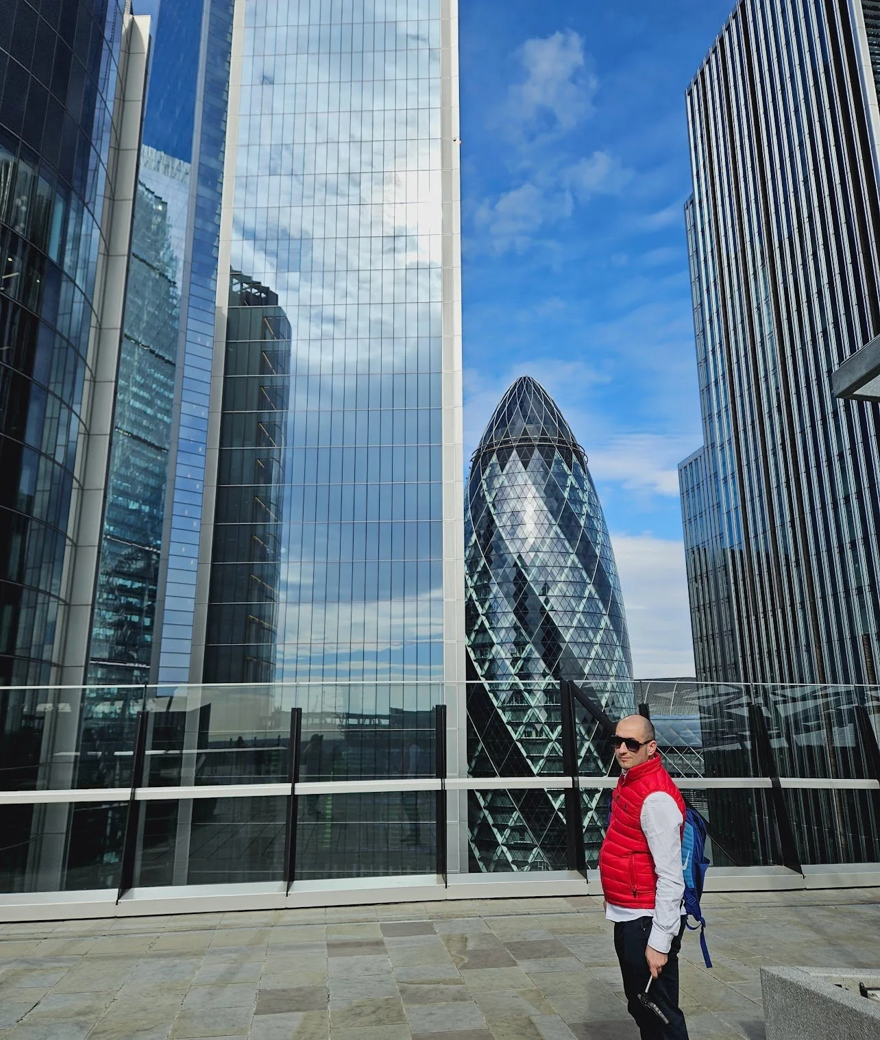 A man wearing sunglasses, a red vest, and carrying a backpack stands in front of tall modern glass buildings with a partly cloudy sky overhead.