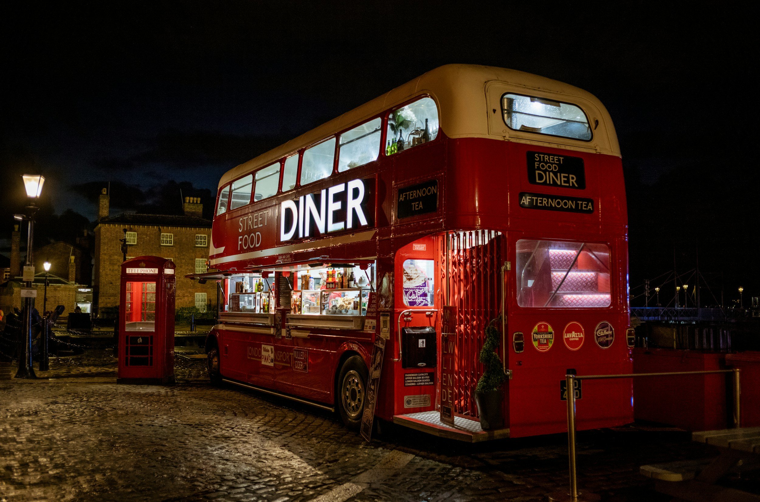 Red double-decker bus serving as street food and diner at night, with signage for street food diner and afternoon tea, parked on cobblestone street near a red telephone booth.