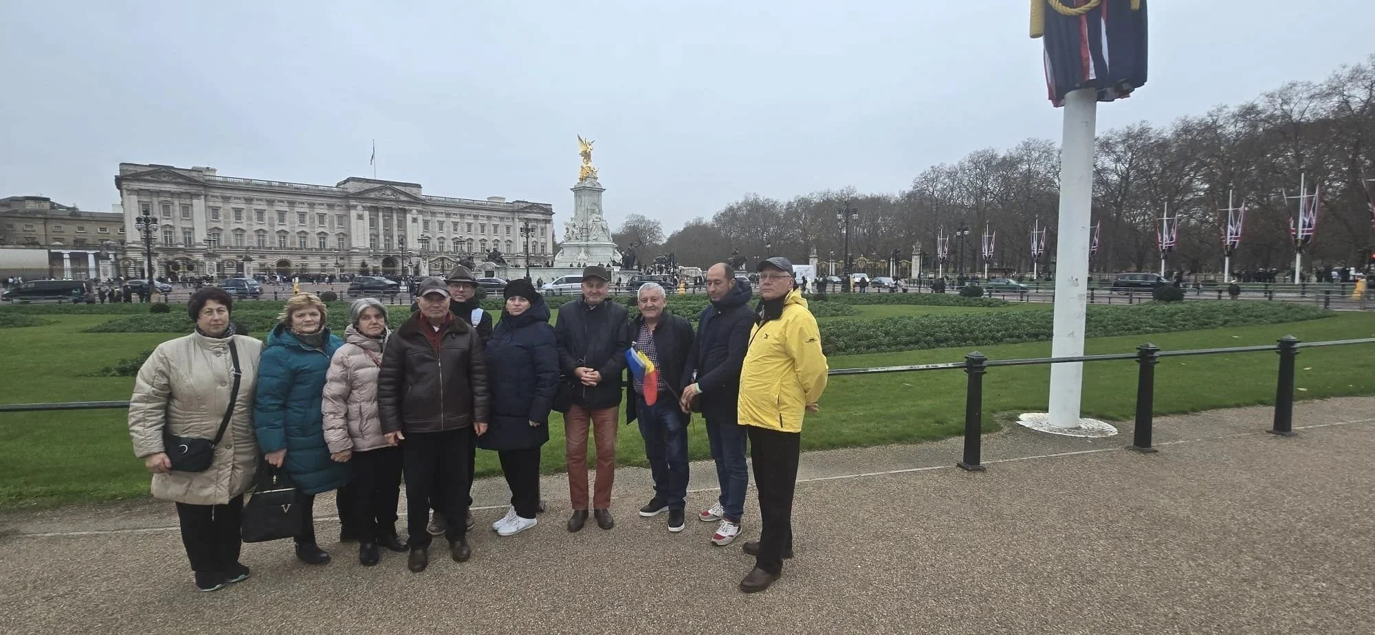 Group of nine people standing in front of Buckingham Palace in London, with a statue and flags visible in the background on a cloudy day.