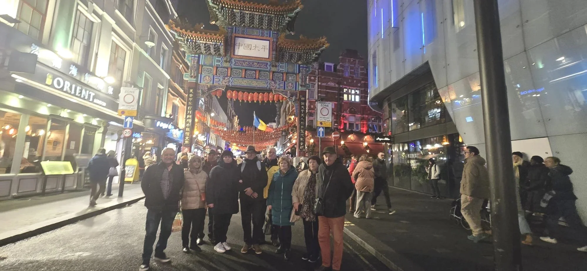 People walking and gathering in a busy street at night near a large Chinese-style gateway decorated with lanterns and traditional architectural details, with shops and modern buildings on either side.
