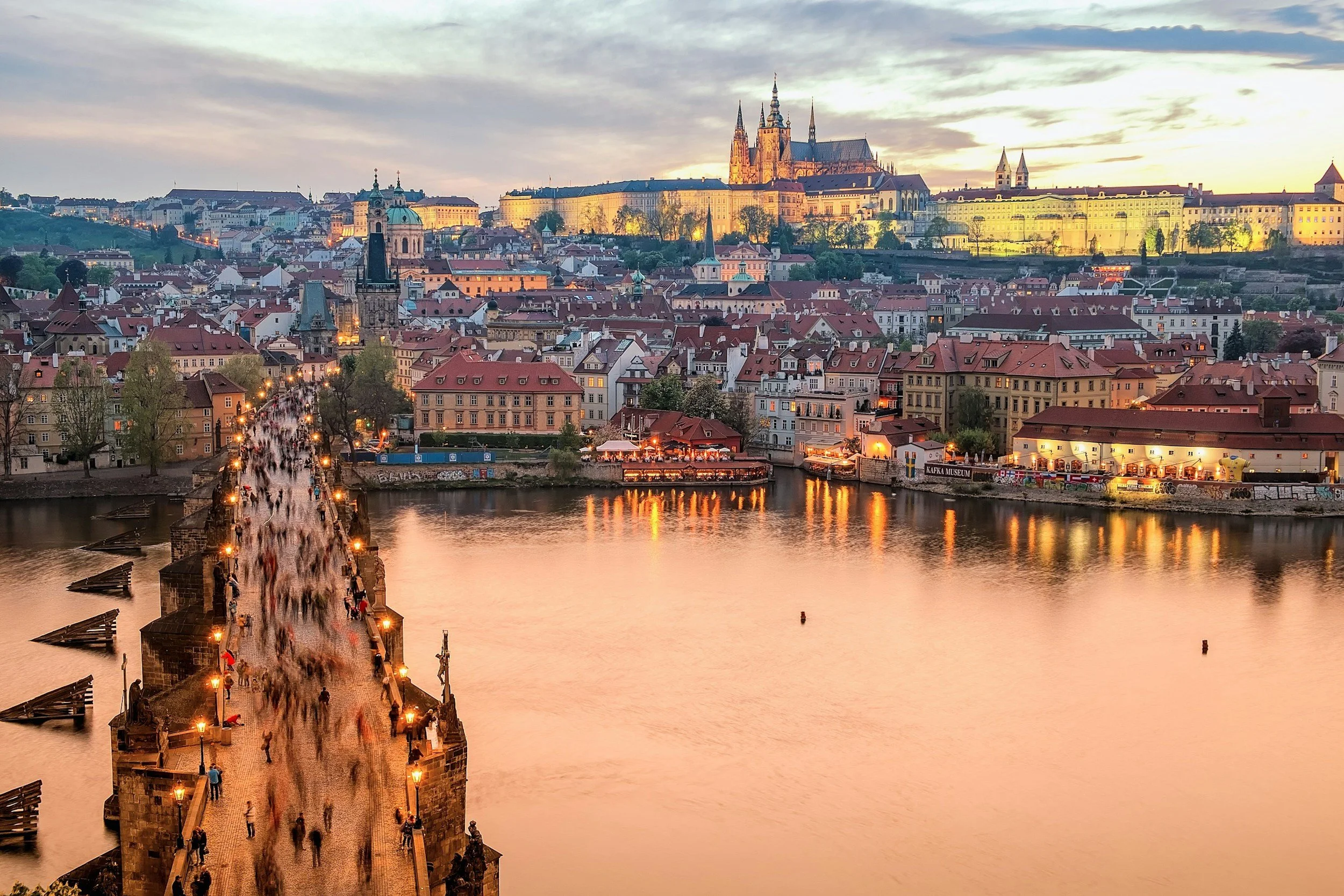Panoramic view of Prague, Czech Republic, during sunset, featuring Charles Bridge with pedestrians, the Vltava River, historic buildings, and Prague Castle in the background.