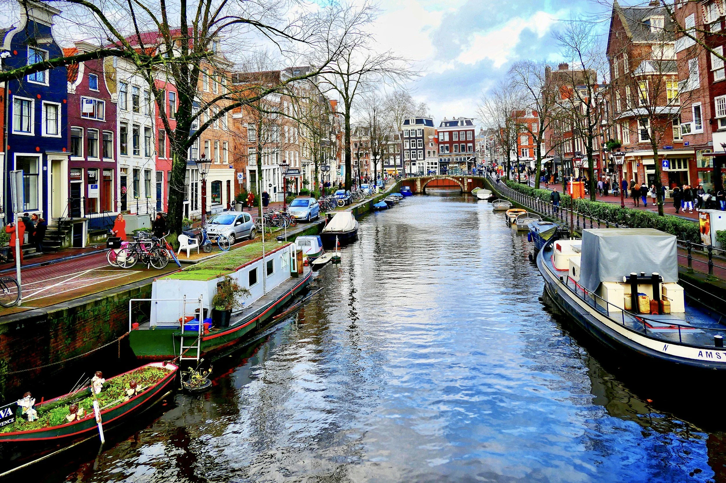 Colorful row of houses along a canal with boats and people walking on the sidewalk in Amsterdam, Netherlands.