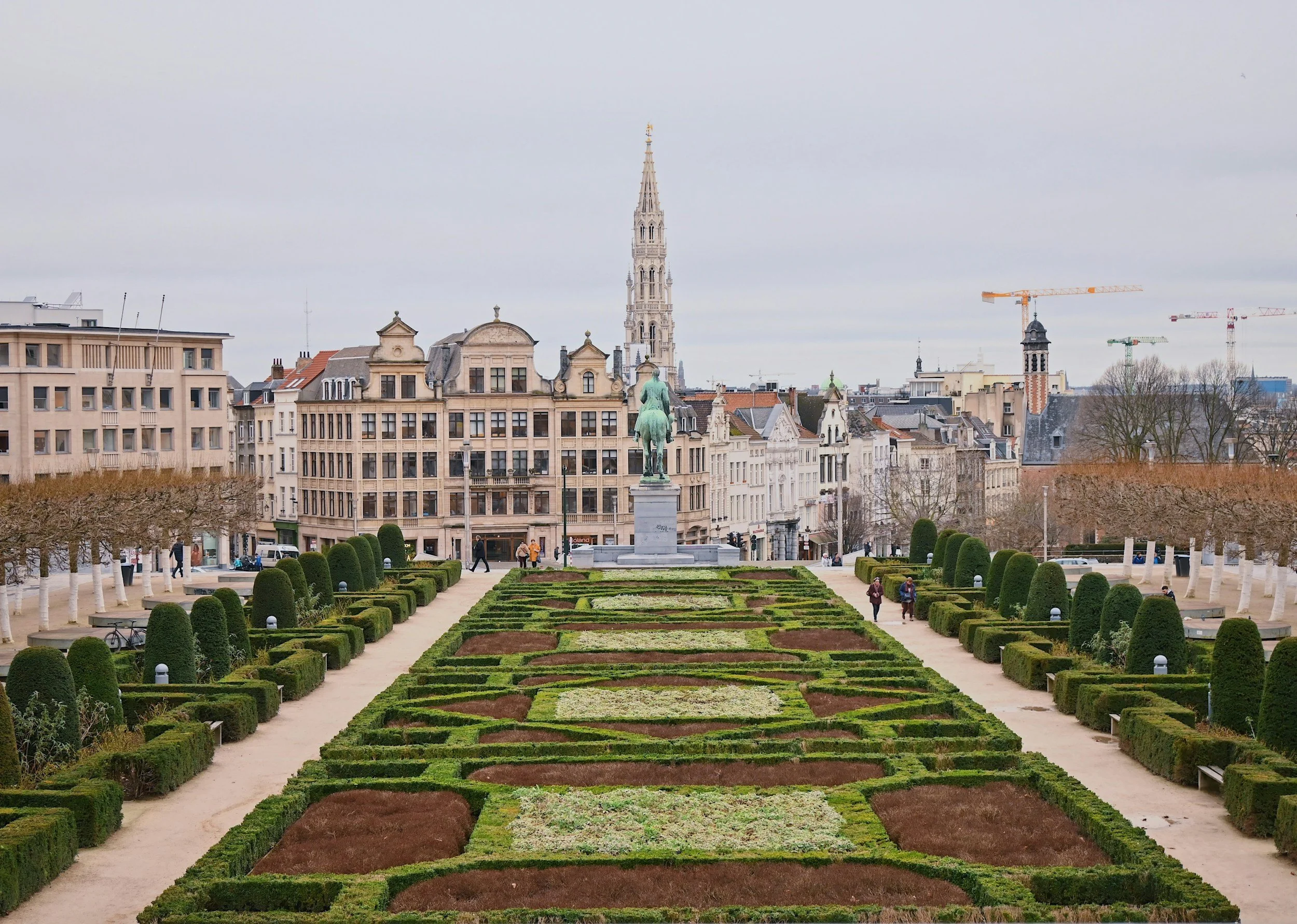 City park with manicured flower beds, trees with white trunks, and a statue of a man on horseback in the center, with historic buildings and church steeples in the background.