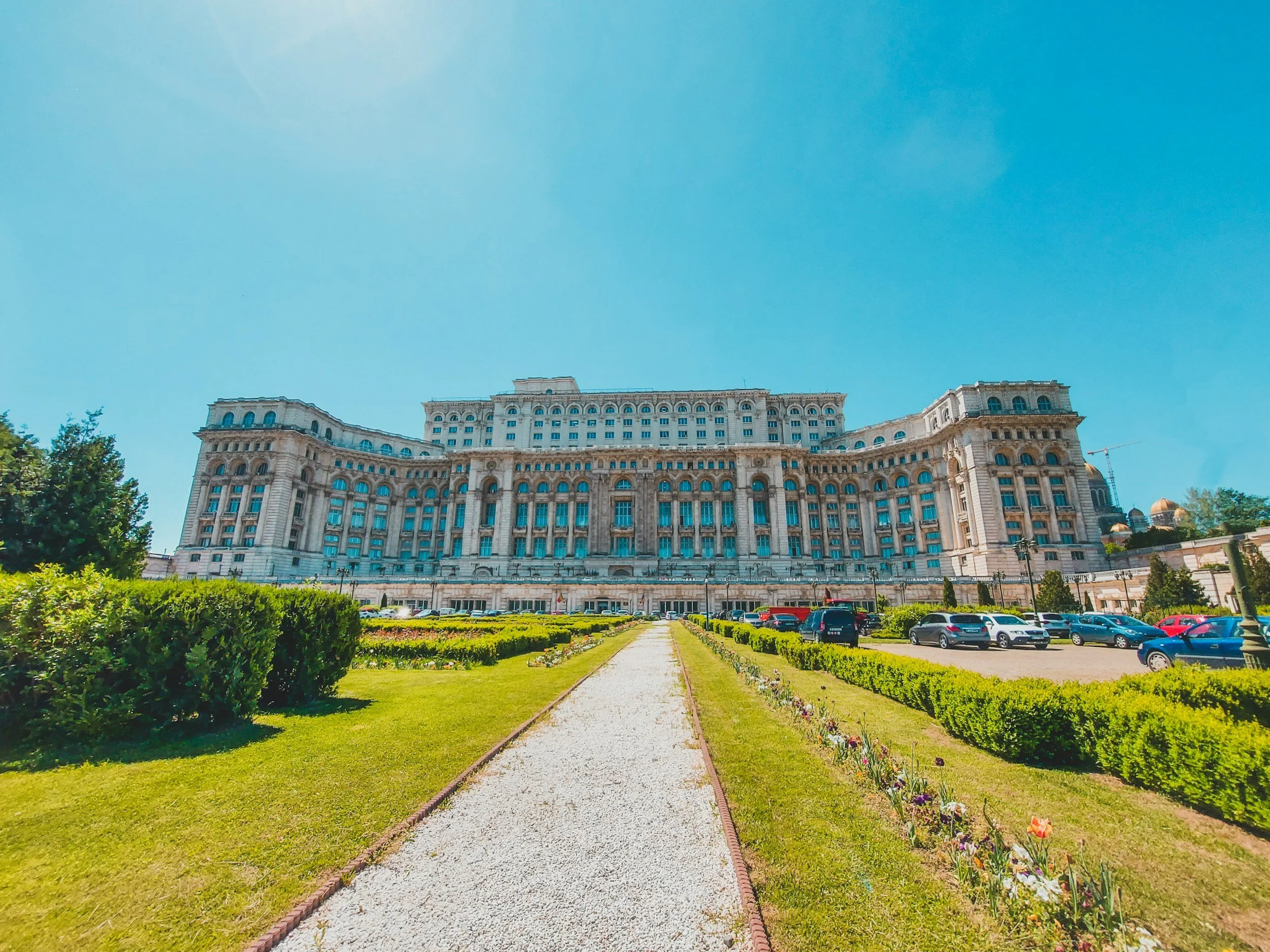Large historic hotel building with multiple floors, ornate architecture, and a symmetrical facade, set against a bright blue sky, with a pathway, green bushes, and parking lot in the foreground.