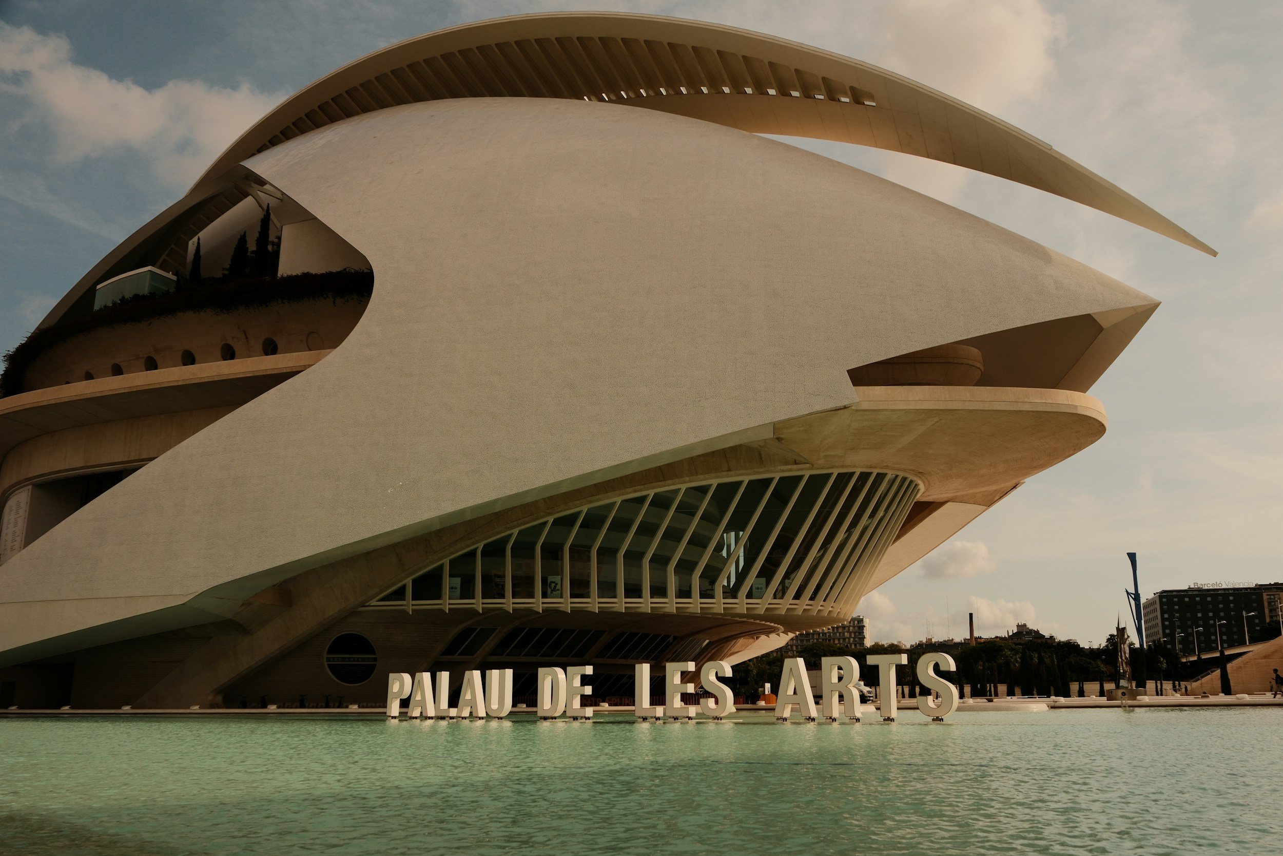 Modern building with curved white structure, part of Palau de les Arts in Valencia, Spain, surrounded by water and with large white letters spelling 'PALAU DE LES ARTS' in front.