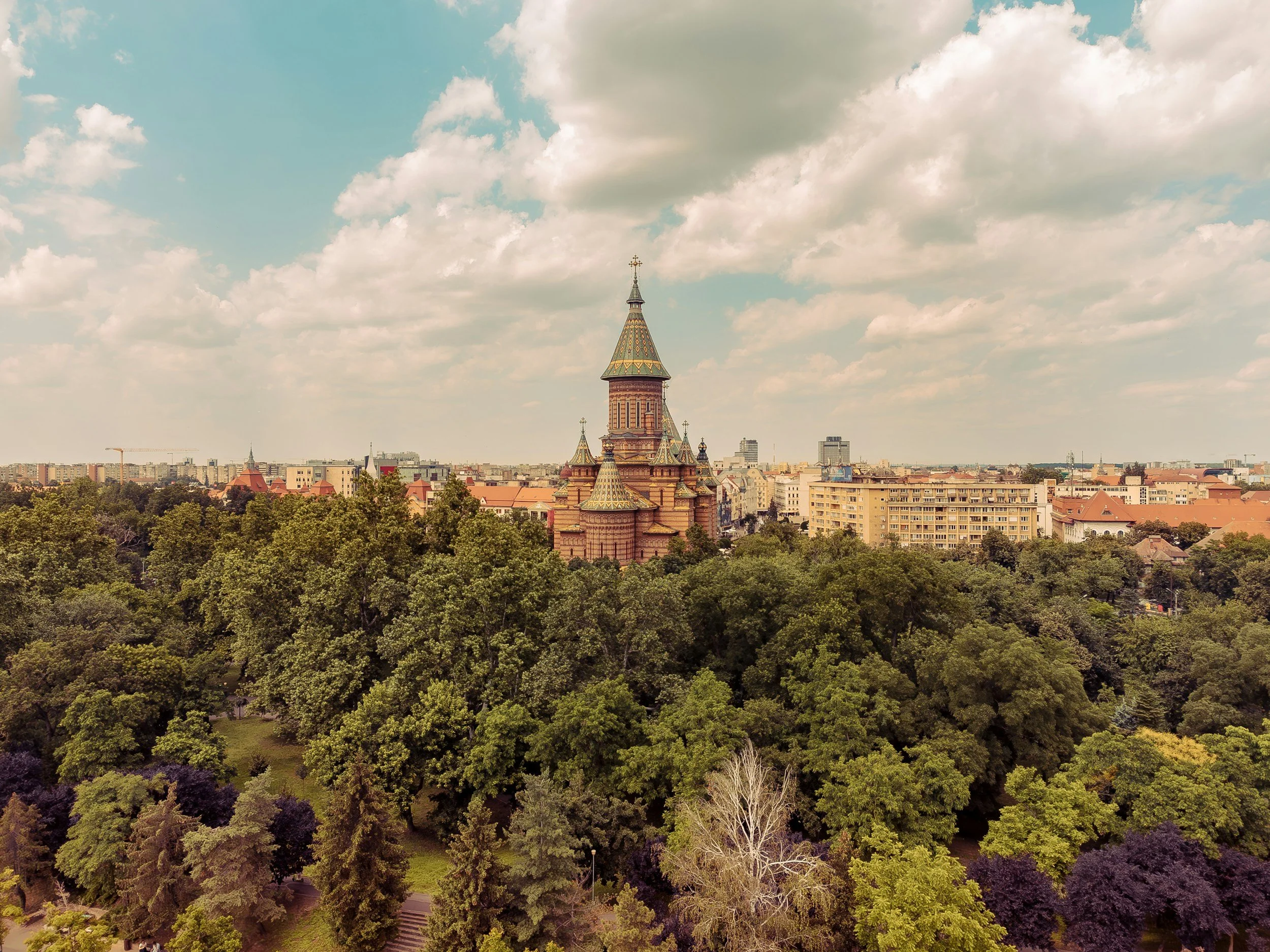 A historic church with a tall, pointed tower surrounded by lush green trees in a cityscape under a partly cloudy sky.