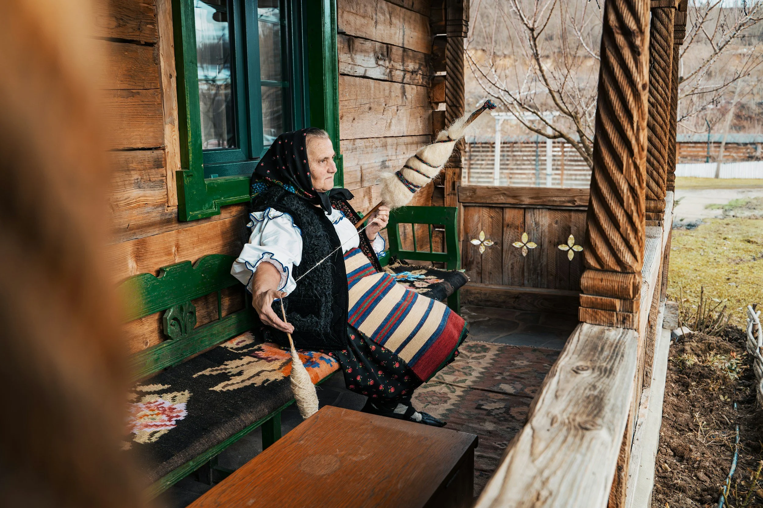 An elderly woman sitting on a decorated wooden bench on a porch, holding traditional spinning tools, dressed in folk attire with a headscarf, in front of a rustic wooden house.