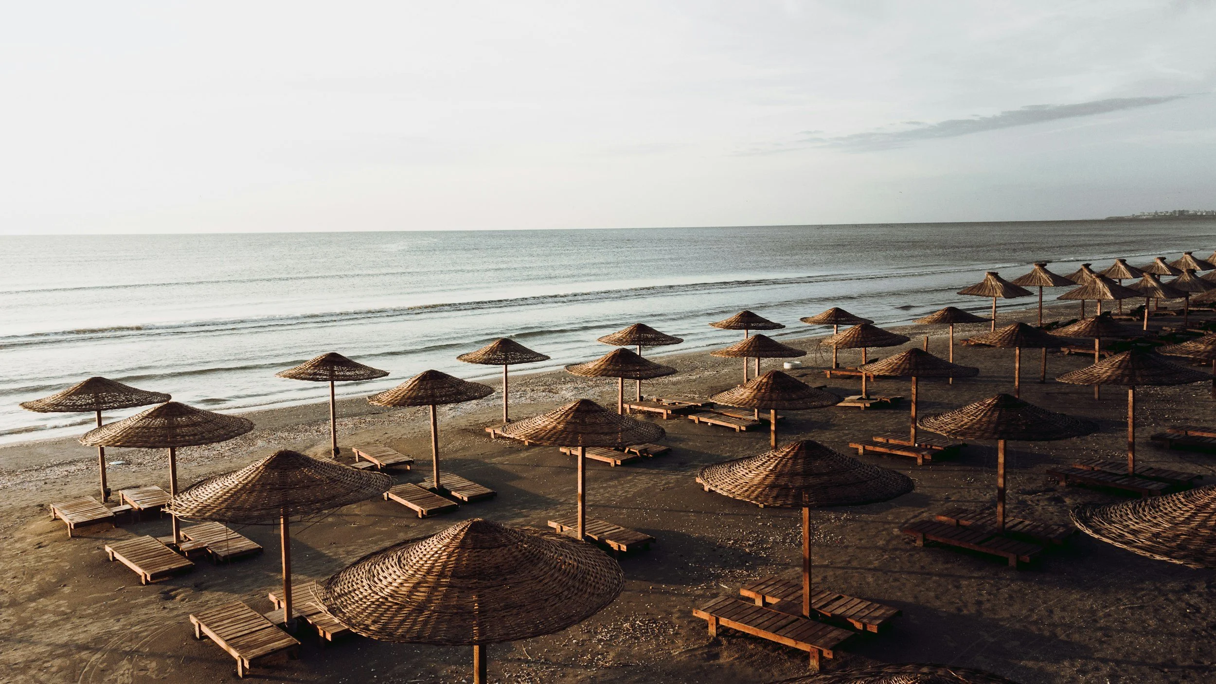 Empty beach with rows of brown straw umbrellas and sun loungers, calm ocean waves, and cloudy sky.