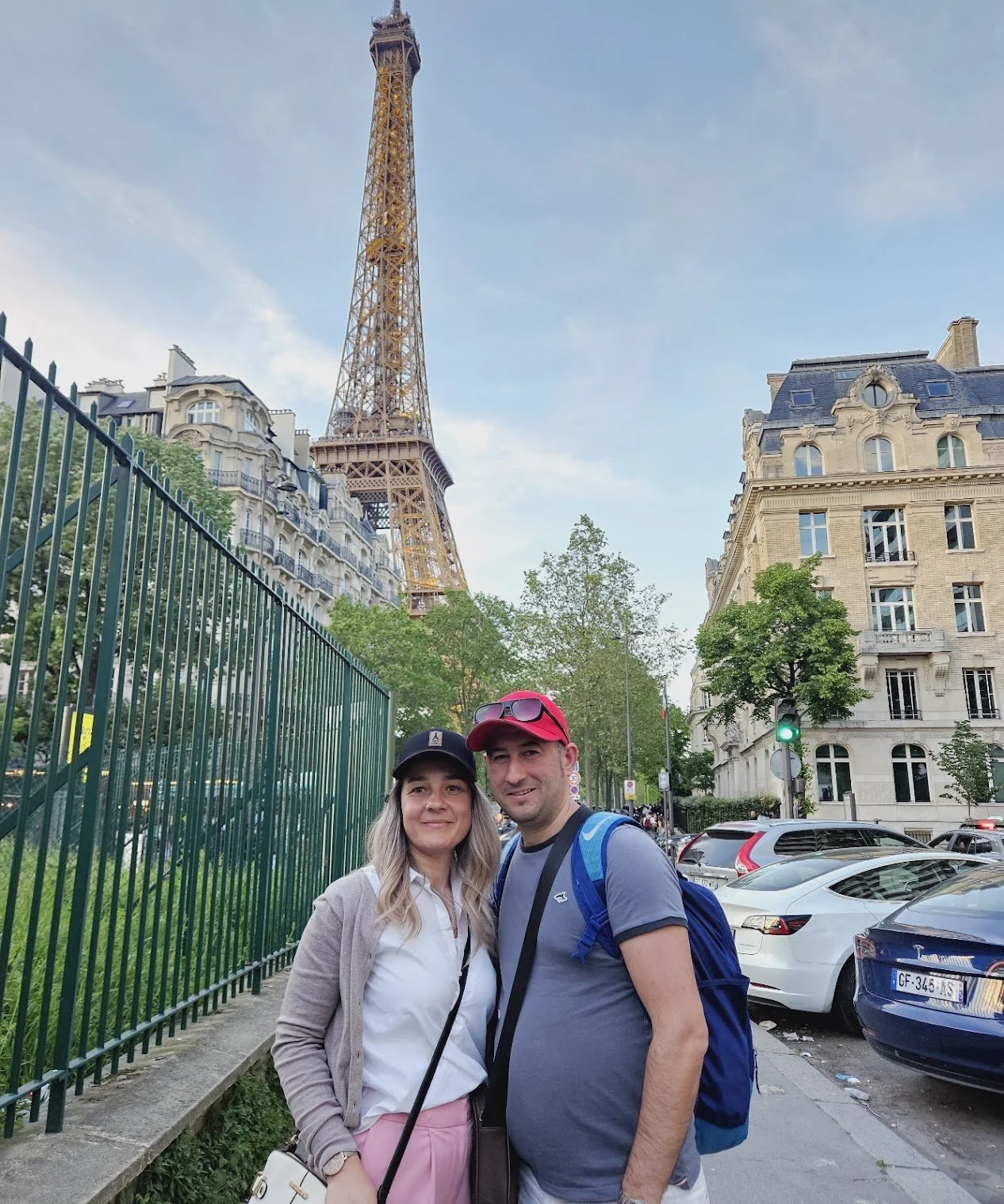 A man and woman standing on a city street with the Eiffel Tower in the background. The woman is wearing a white shirt, pink pants, a black cap, and has blonde hair. The man is wearing a gray t-shirt, a red cap, and has a blue backpack. There are park