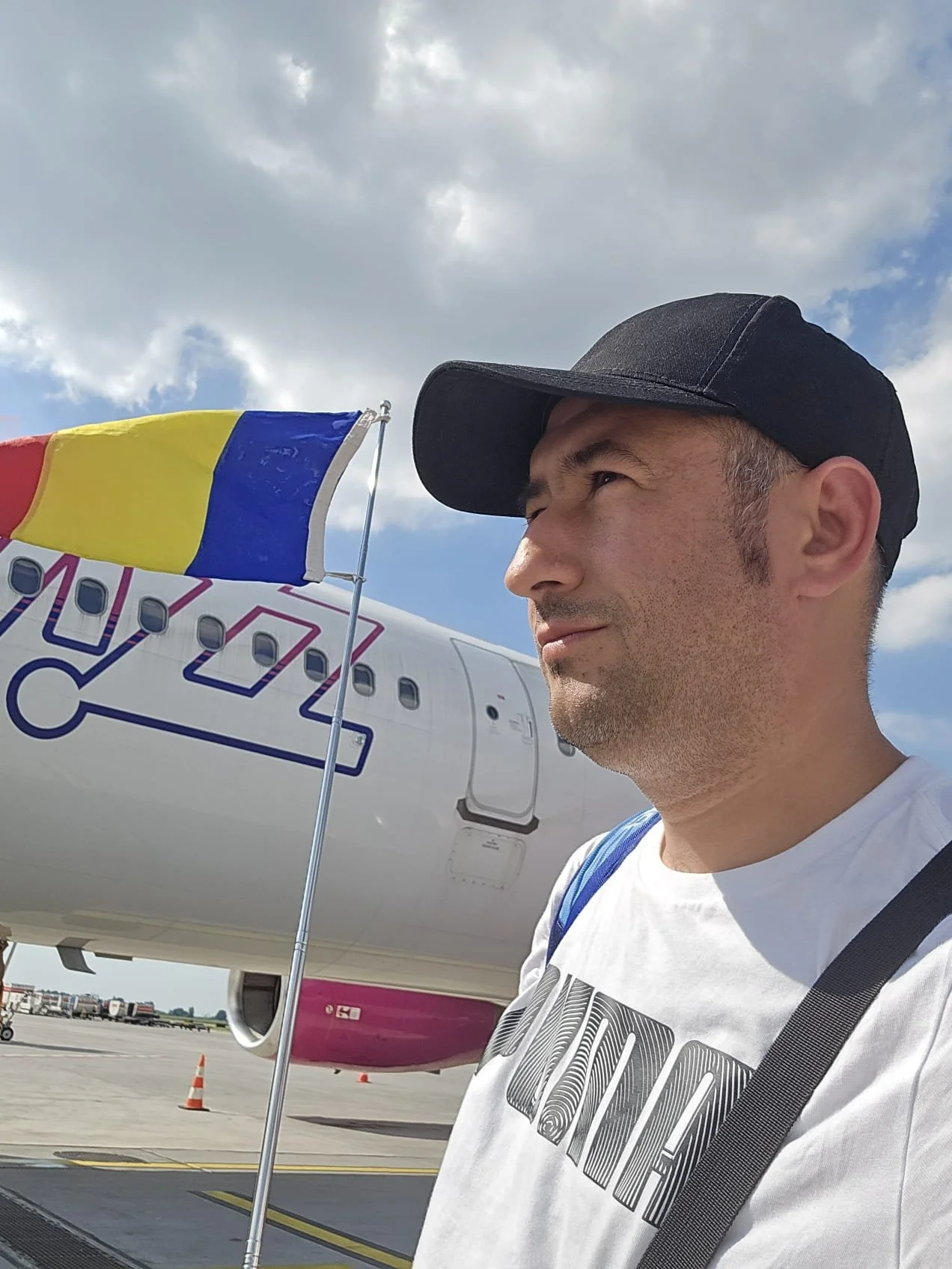 Man at an airport standing near an airplane with a Romanian flag in front of him, with a cloudy sky overhead.