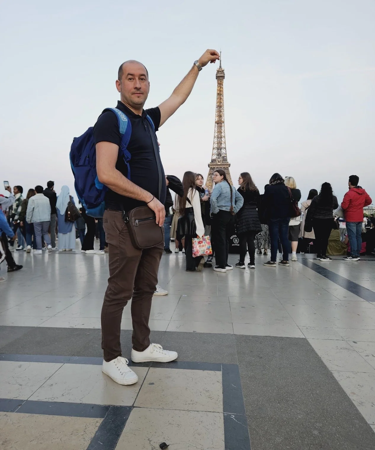 A man standing in front of the Eiffel Tower in Paris, France, holding a miniature Eiffel Tower replica, with a crowd of tourists in the background.