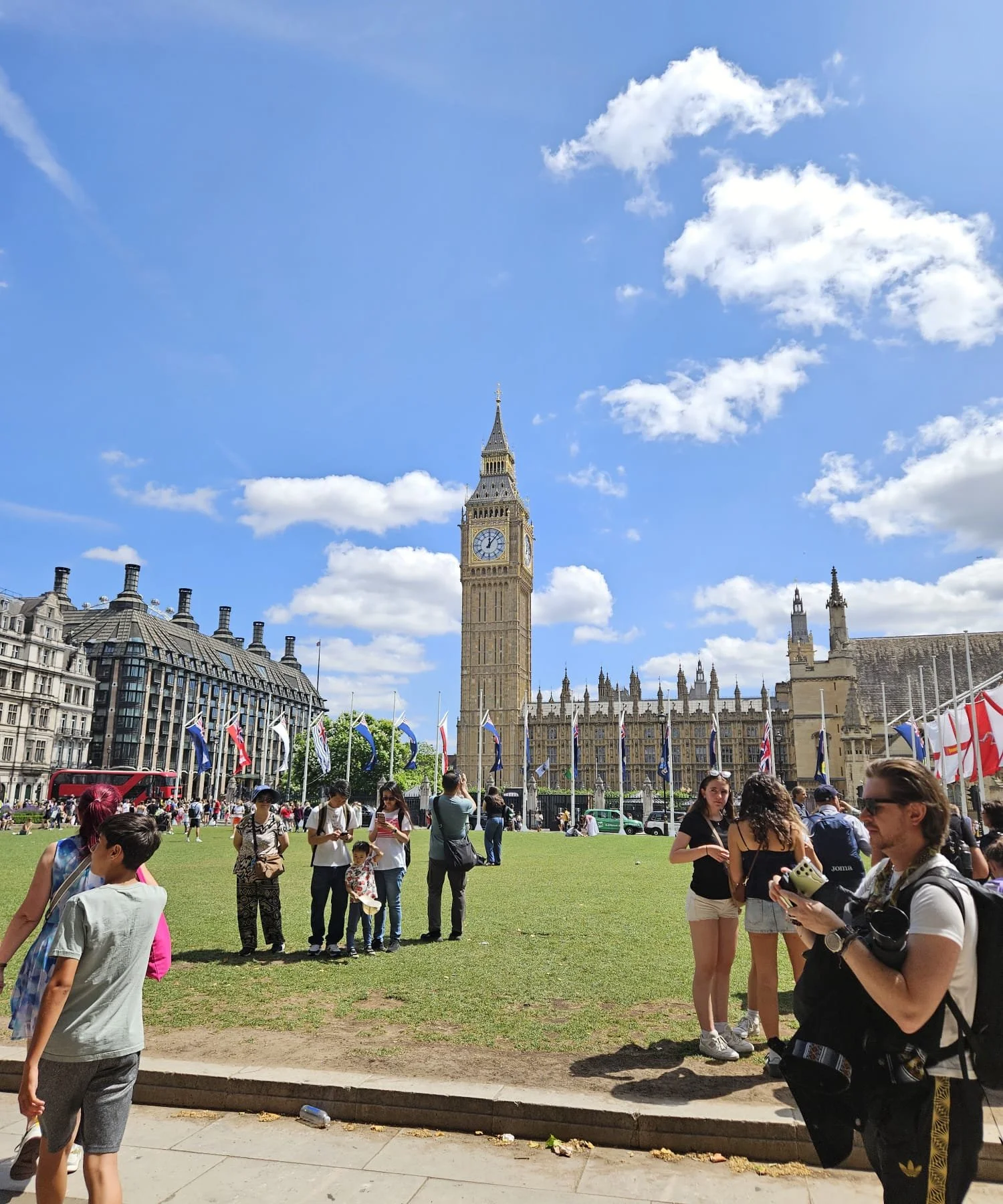 People gather on a grassy area near Big Ben in London, with historic buildings and flags in the background, under a partly cloudy sky.