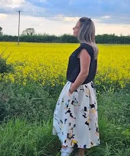 Woman standing in a field of yellow flowers, with trees and a blue sky in the background.