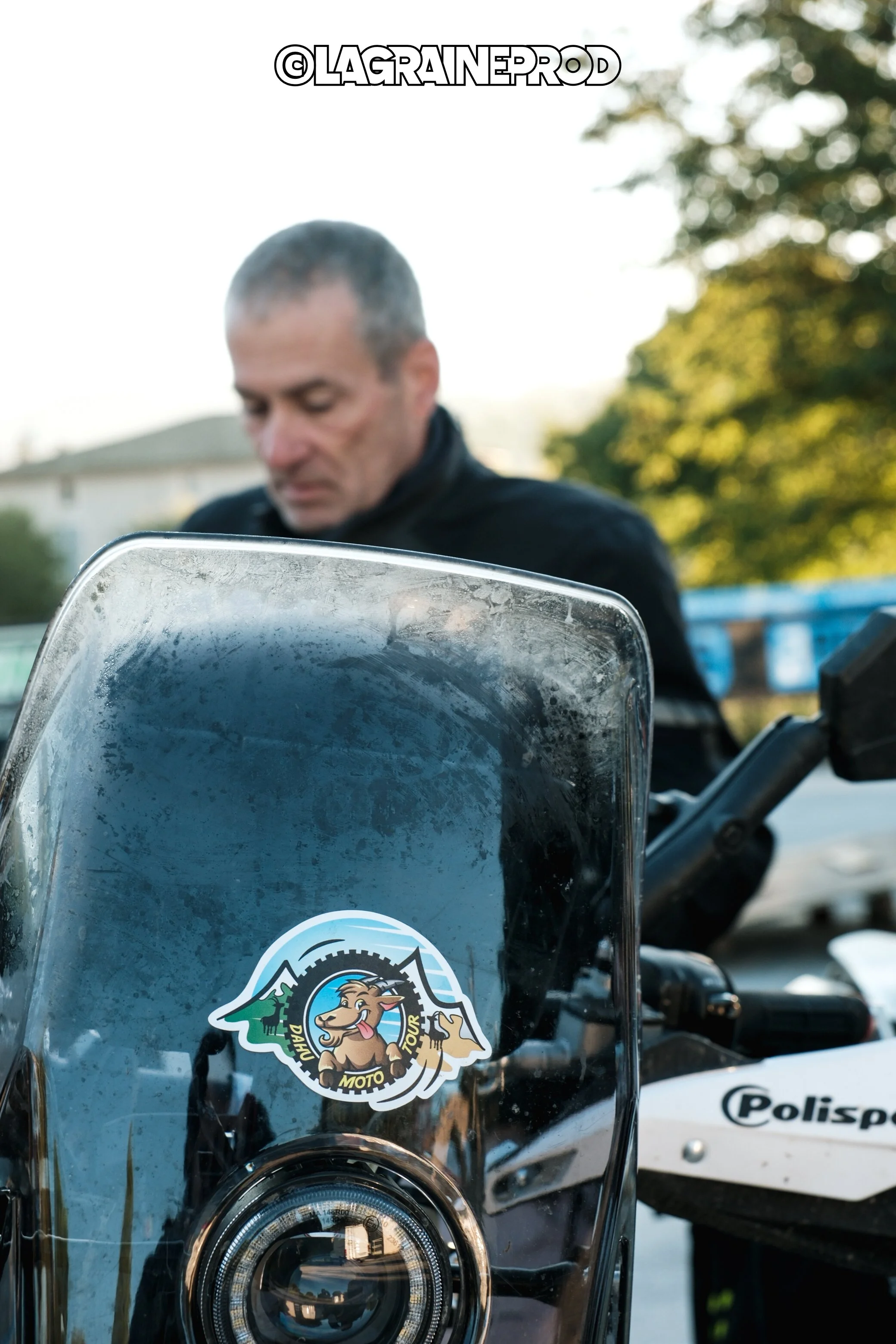 Un homme debout près d'une moto avec un pare-brise transparent portant un autocollant illustrant un chien avec un paysage de montagne, une boule à neige et des pièces de monnaie.