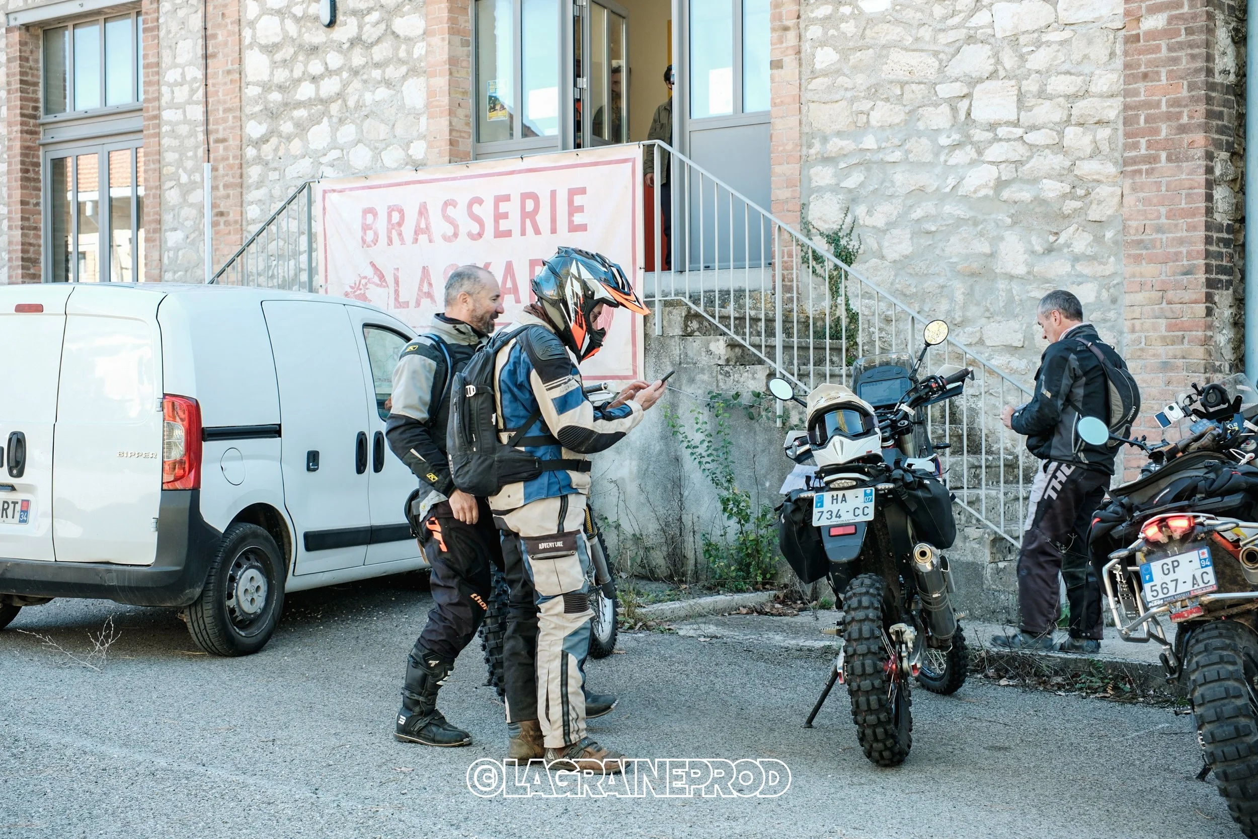Groupe de motards habillés de vêtements de protection autour de motos tout-terrain, devant un restaurant "Brasserie La Grange" avec un bâtiment en pierre en arrière-plan.