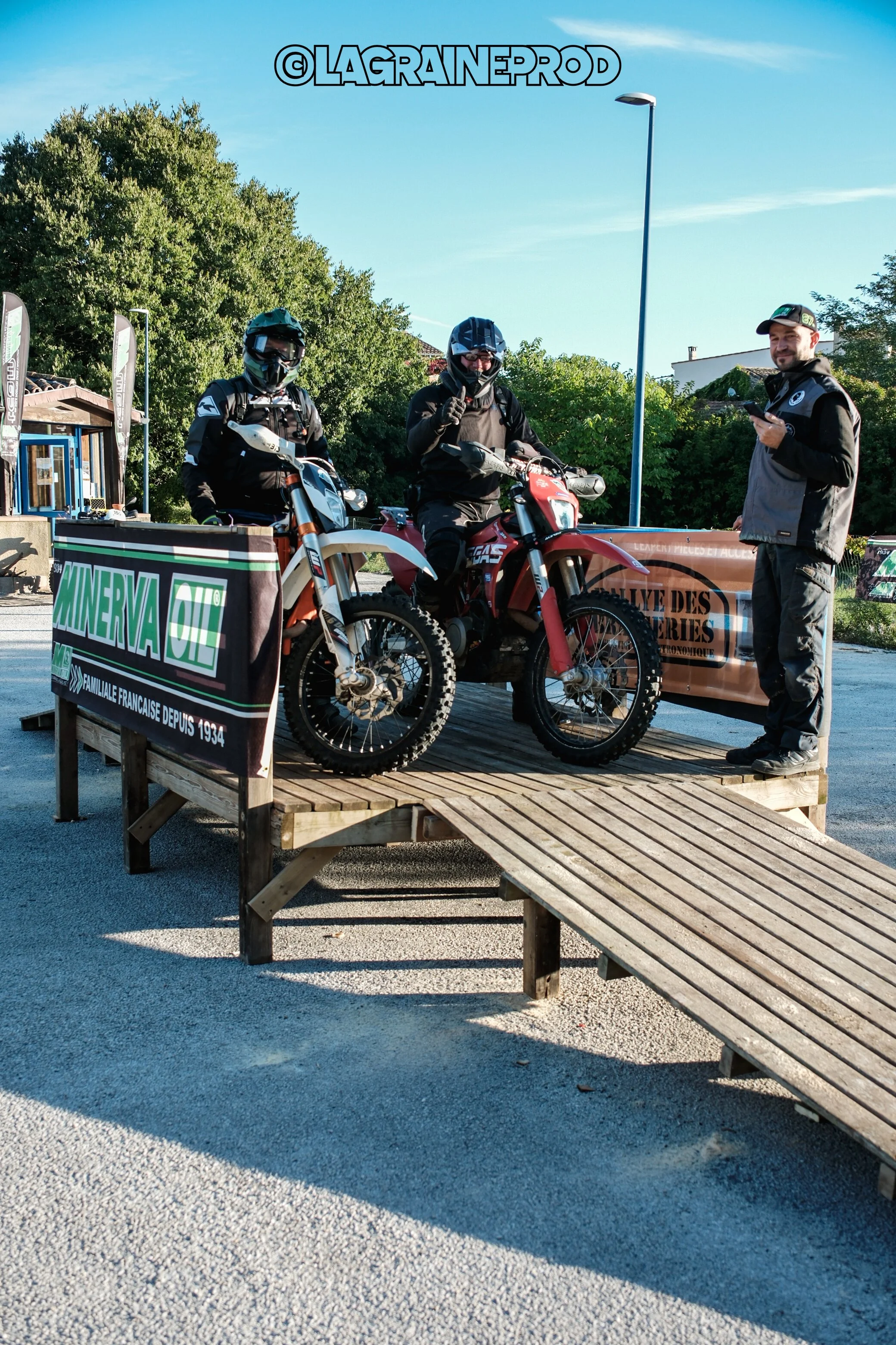 Deux motards en tenue de randonnée sur des motos tout-terrain, avec un homme debout à côté d'eux qui regarde son téléphone, sur une plateforme en bois, avec des panneaux publicitaires en arrière-plan et un ciel clair.