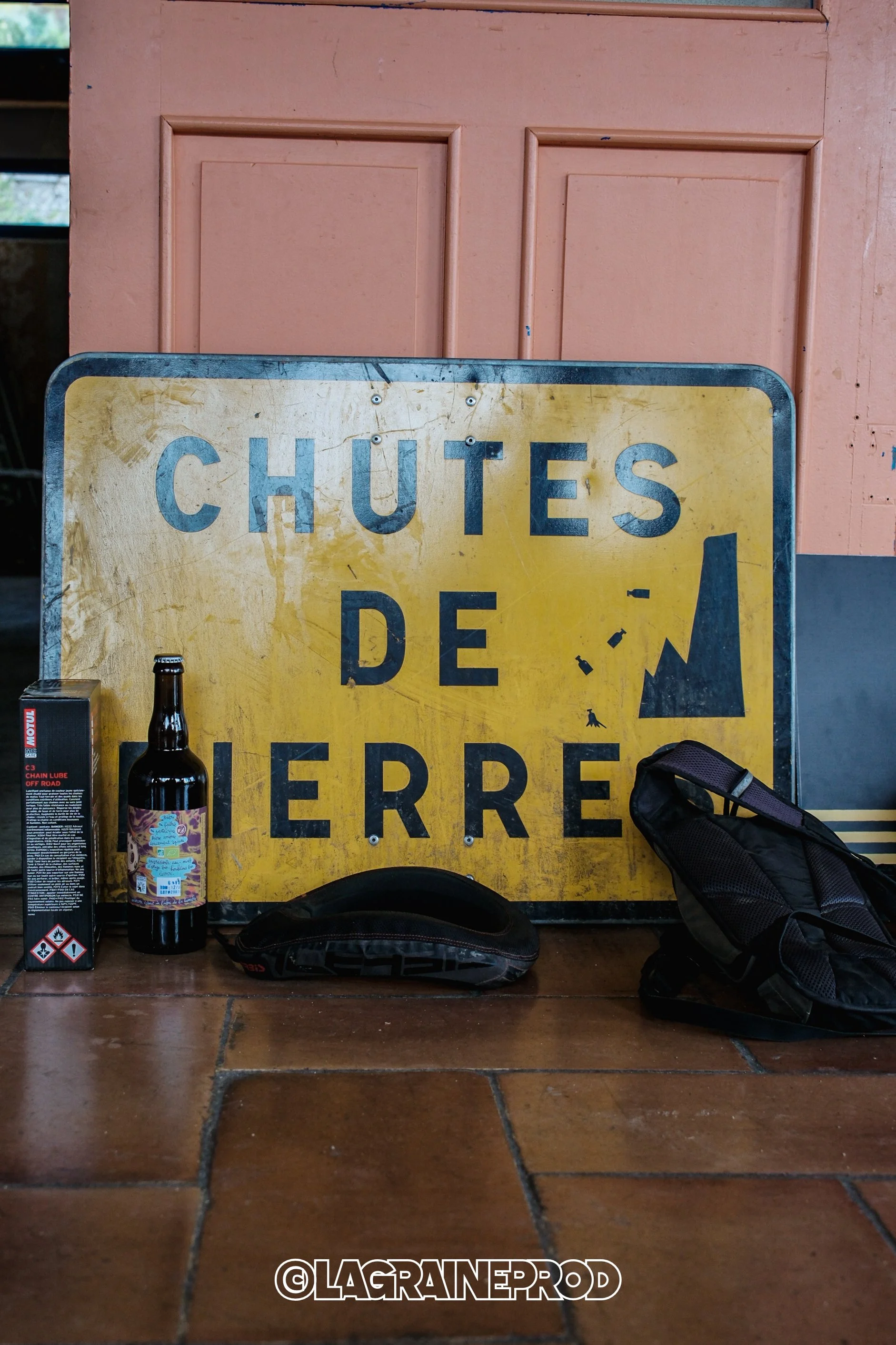 Une pancarte jaune avec des lettres bleues disant 'Chutes de terre', accompagnée d'une bouteille, d'une boîte de lubrifiant, d'une paire de gants et d'un sac à dos, posés sur un sol en carrelage en face d'une porte en bois rose.
