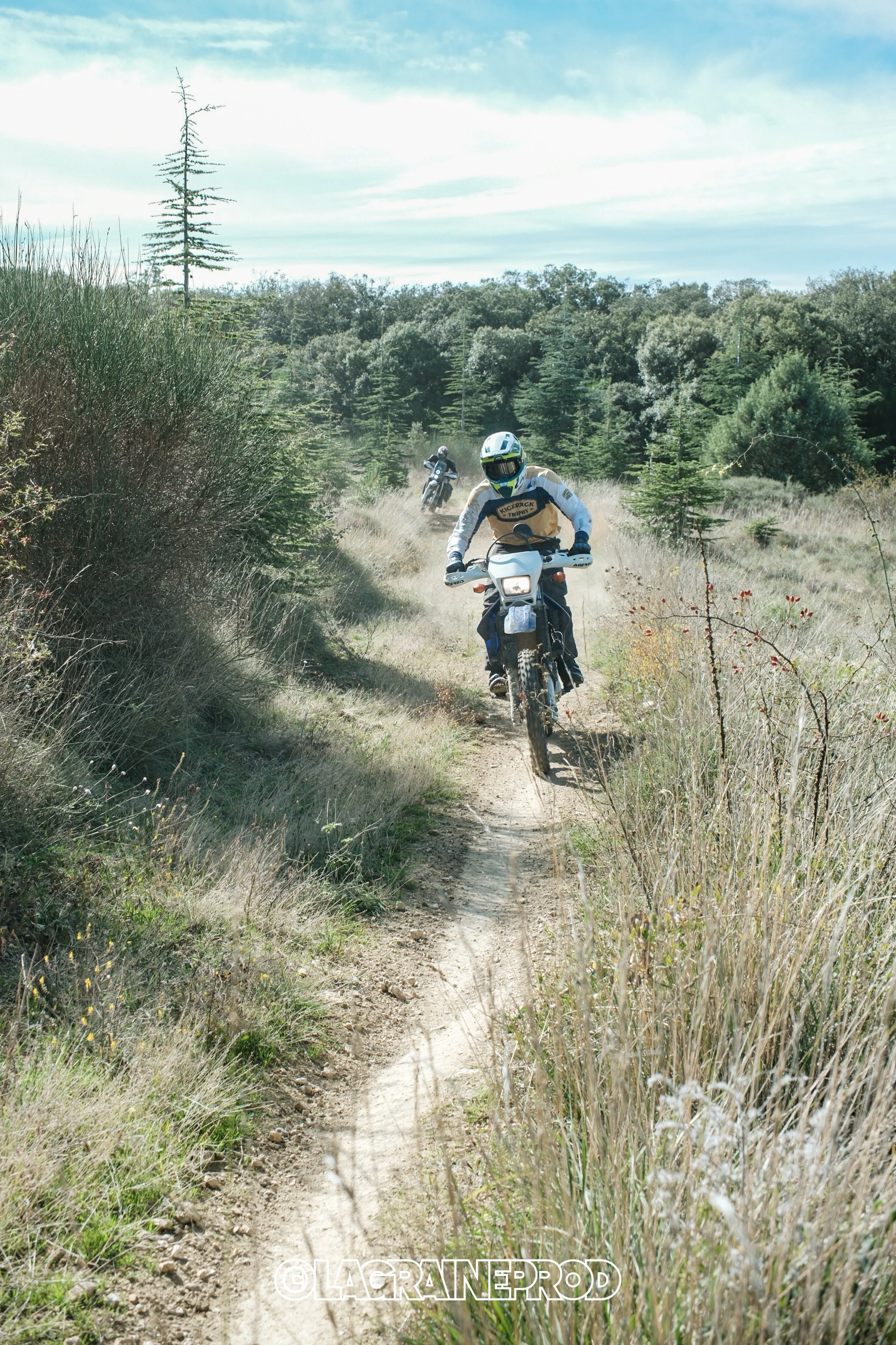 Deux motocyclistes traversent un sentier étroit à travers une zone herbeuse et boisée en plein air.